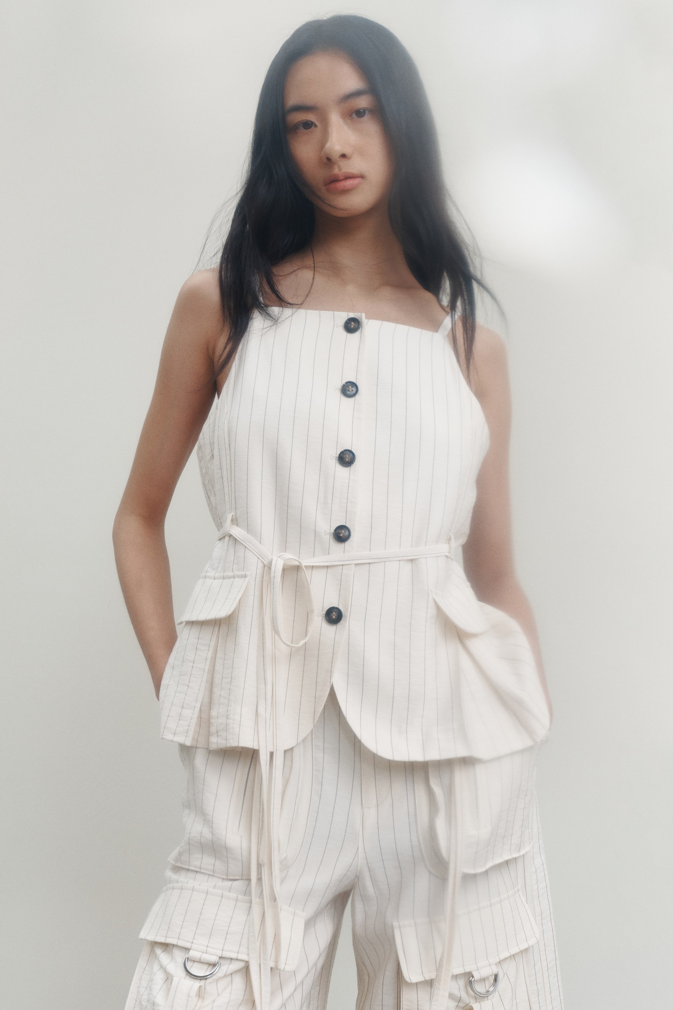View larger image: A light-colored pinstriped sleeveless top dan seluar pendek kargo yang sepadan sedang dipamerkan dari hadapan. Top ini menampilkan leher berbentuk segi empat, penutupan butang di hadapan dengan lima butang gelap, tali fabrik yang mengikat pinggang, dan dua poket flap bulat. Seluar pendek kargo ini juga menampilkan pelbagai poket tampalan, beberapa termasuk D-rings, dan menunjukkan tali fabrik yang tergantung.