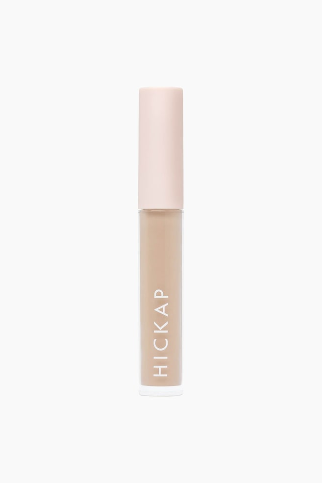 Et cylindrisk rør med flydende kosmetik står oprejst og vender direkte fremad. Den gennemsigtige nederste del af røret viser en lys beige væske, mens en uigennemsigtig lyserød hætte dækker toppen. Mærkenavnet "HICKAP" er trykt lodret med hvide bogstaver på den klare krop. - 1