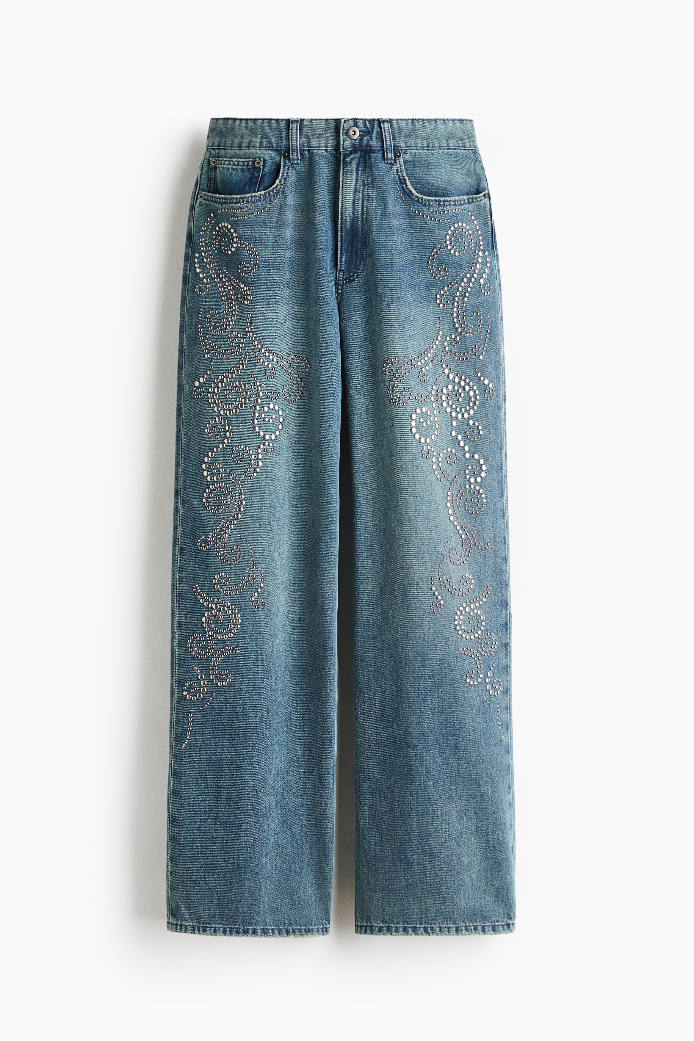 La modelo lleva unos jeans de mezclilla azul con adornos de pedrerÃ­a a lo largo de los costados exteriores de las piernas rectas. Los jeans se combinan con un cinturÃ³n negro en la cintura y una camisa blanca crema bordada con botones, metida dentro de la pretina.