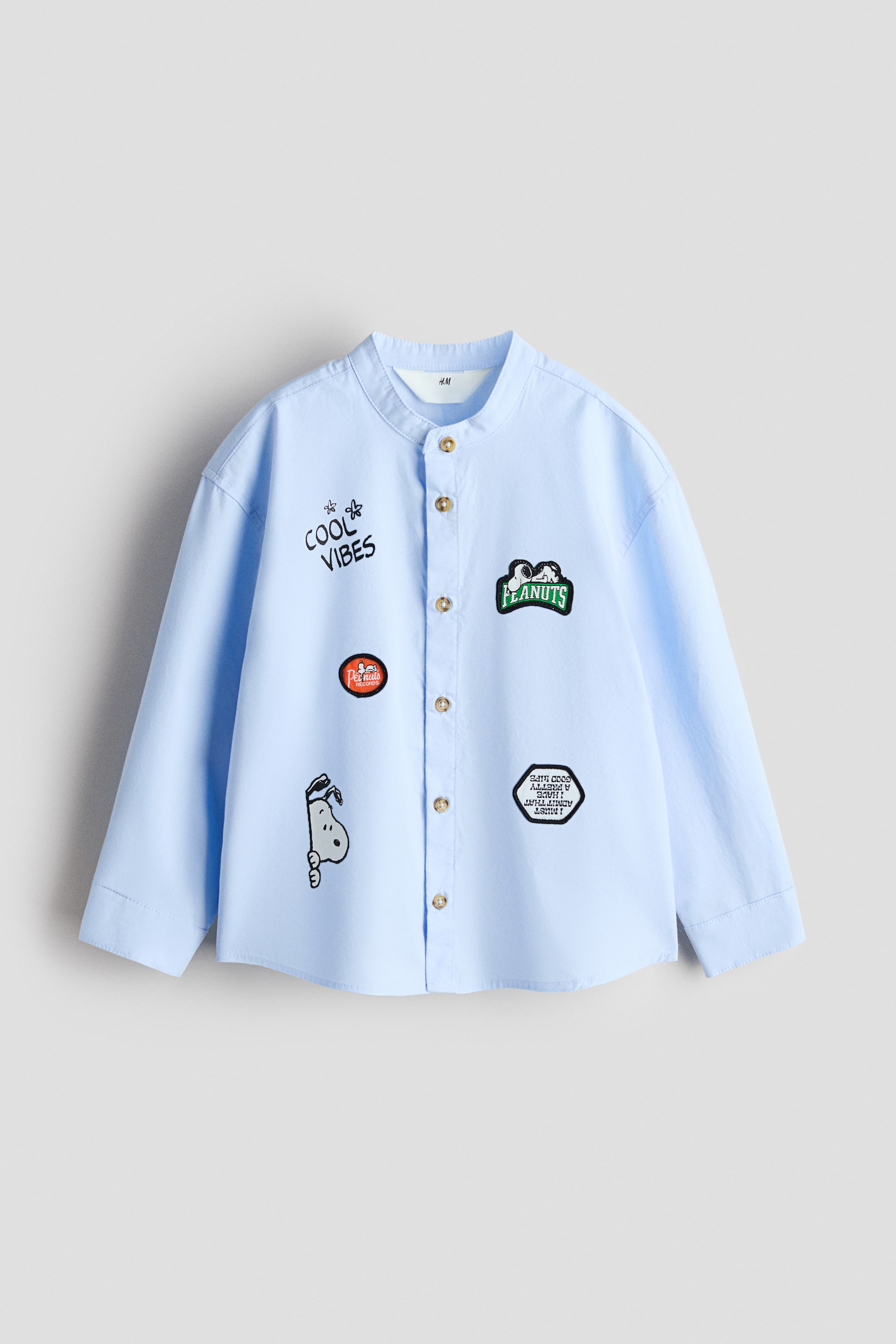 拡大画像を表示: ライトブルーの長袖シャツがFRONT(正面)から撮影されており、バンドカラーと前立てに沿って並んだゴールドカラーのボタンが特徴です。さまざまな刺繍ワッペンで飾られており、"COOL VIBES"、Snoopy と Woodstock が描かれた "PEANUTS"、丸い赤の "Peanuts" ワッペン、Snoopy の頭のモチーフ、白い文字入りの黒い八角形ワッペンなどが付いています。