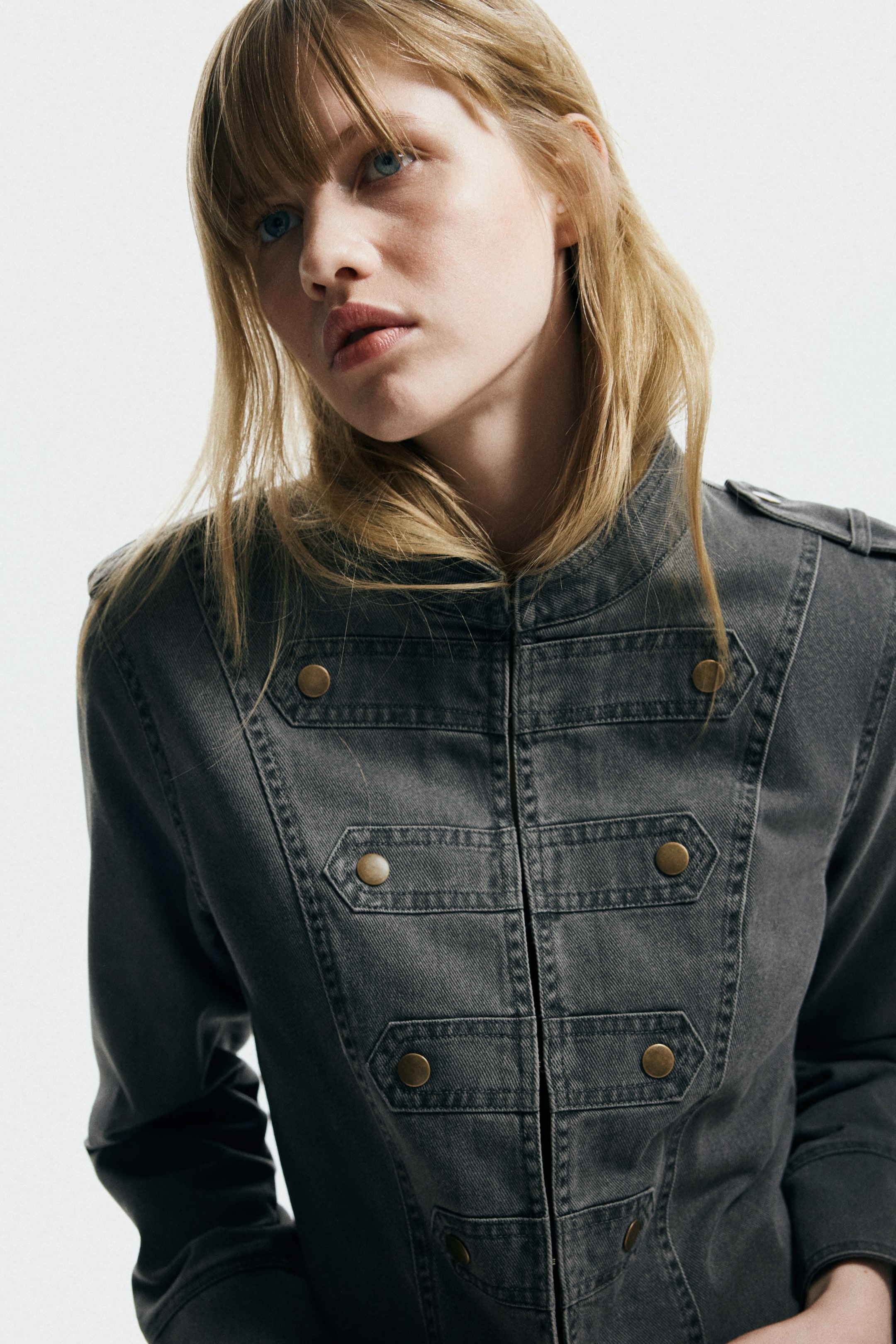 Agrandir l'image: Une veste en denim gris foncé de style militaire, dotée d'un col montant, d'une fermeture éclair centrale à l'avant, de plusieurs paires de boutons décoratifs couleur laiton antique disposés verticalement, et d'épaulettes avec boutons simples.