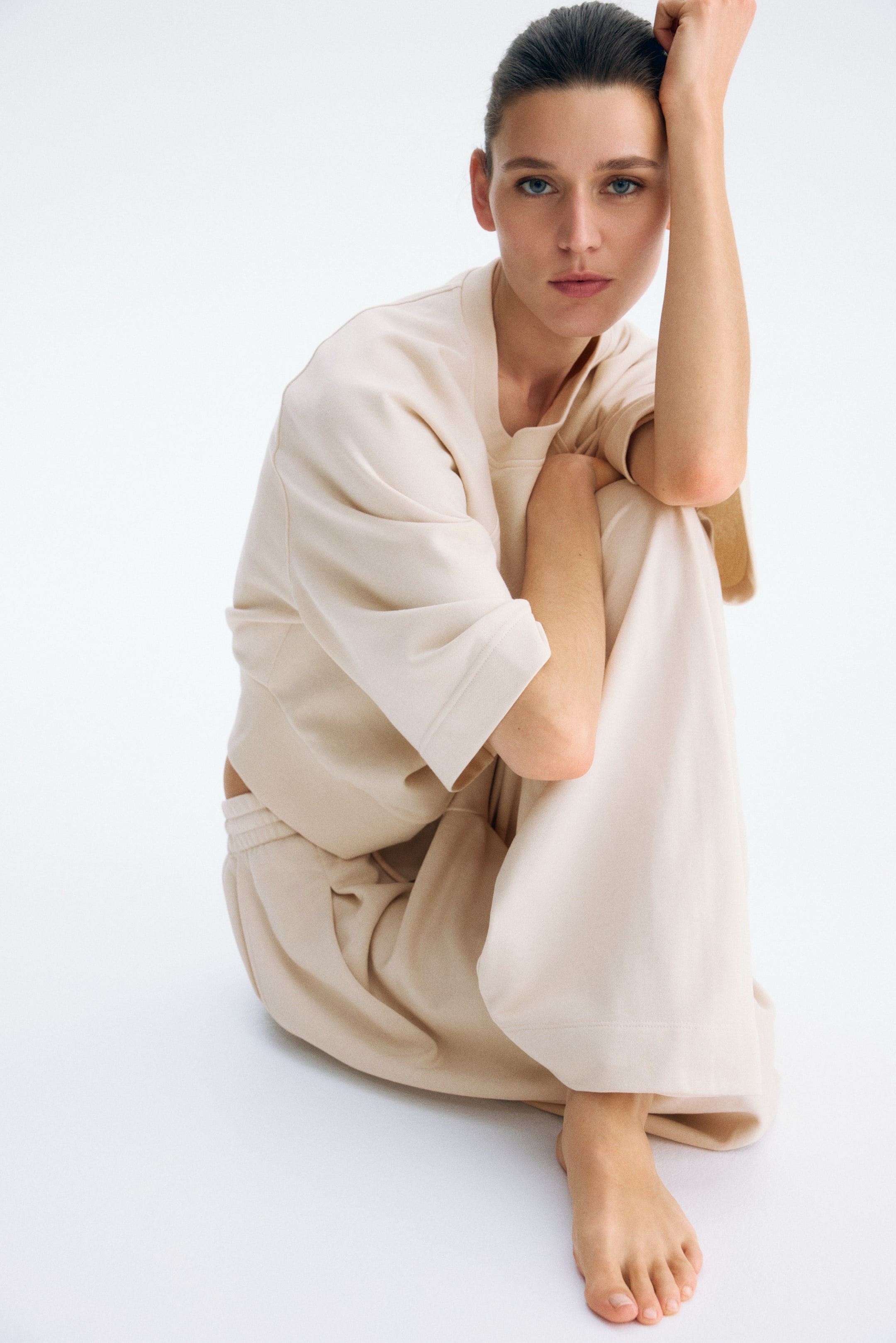 Büyük resmi görüntüle: Açık bej iki parçalı set, rahat kesim, kısa kollu, bisiklet yaka ve geniş kollu, göbek kısmını gösteren bir üst ile öne çıkıyor. Uyumlu geniş paçalı pantolonlar, lastikli bel bandına sahip olup bileklerin etrafında toplanıyor.