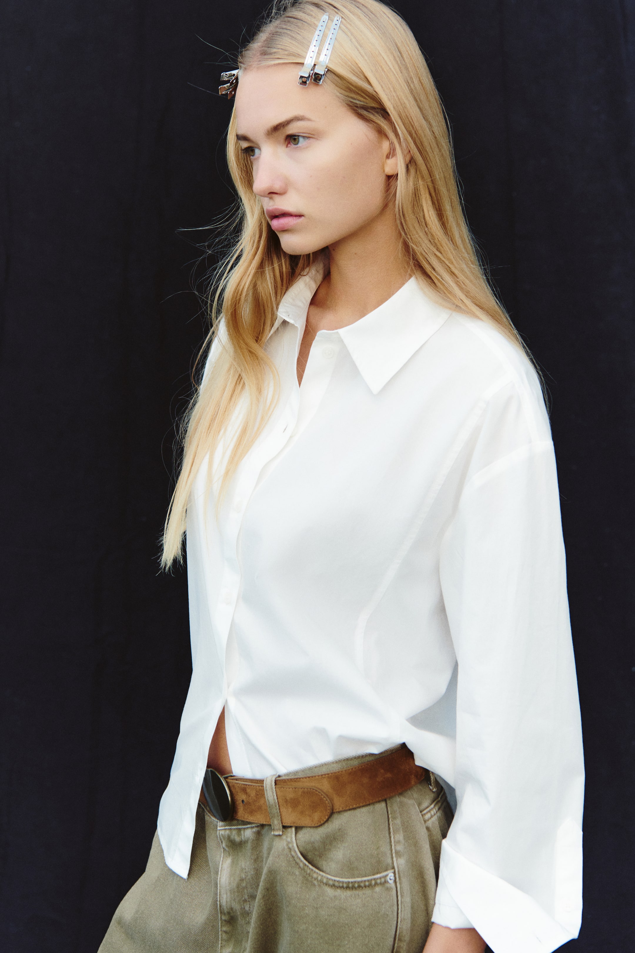 View larger image: A white cotton poplin shirt with a pointed collar and dropped shoulders is worn by the model. The long-sleeve, button-up shirt is styled partially tucked into olive green trousers with a brown belt.