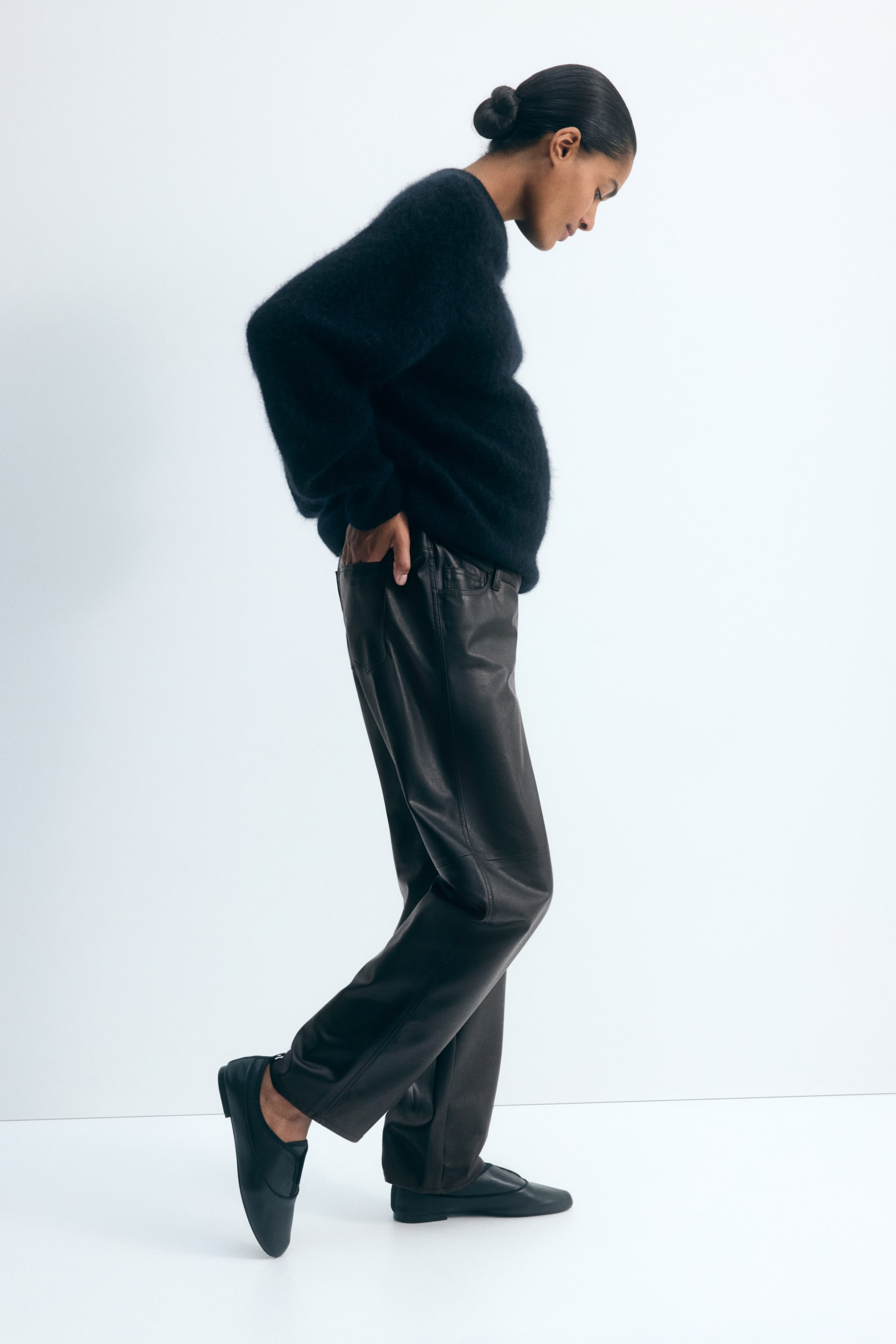 Grotere afbeelding bekijken: De model draagt een donkerbruine broek van gecoat materiaal en staat in profiel met één hand in een achterzak. De broek heeft een losvallend barrel-leg silhouet met een zichtbare horizontale naad ter hoogte van de knie en is gecombineerd met een donker, pluizig gebreid sweater.