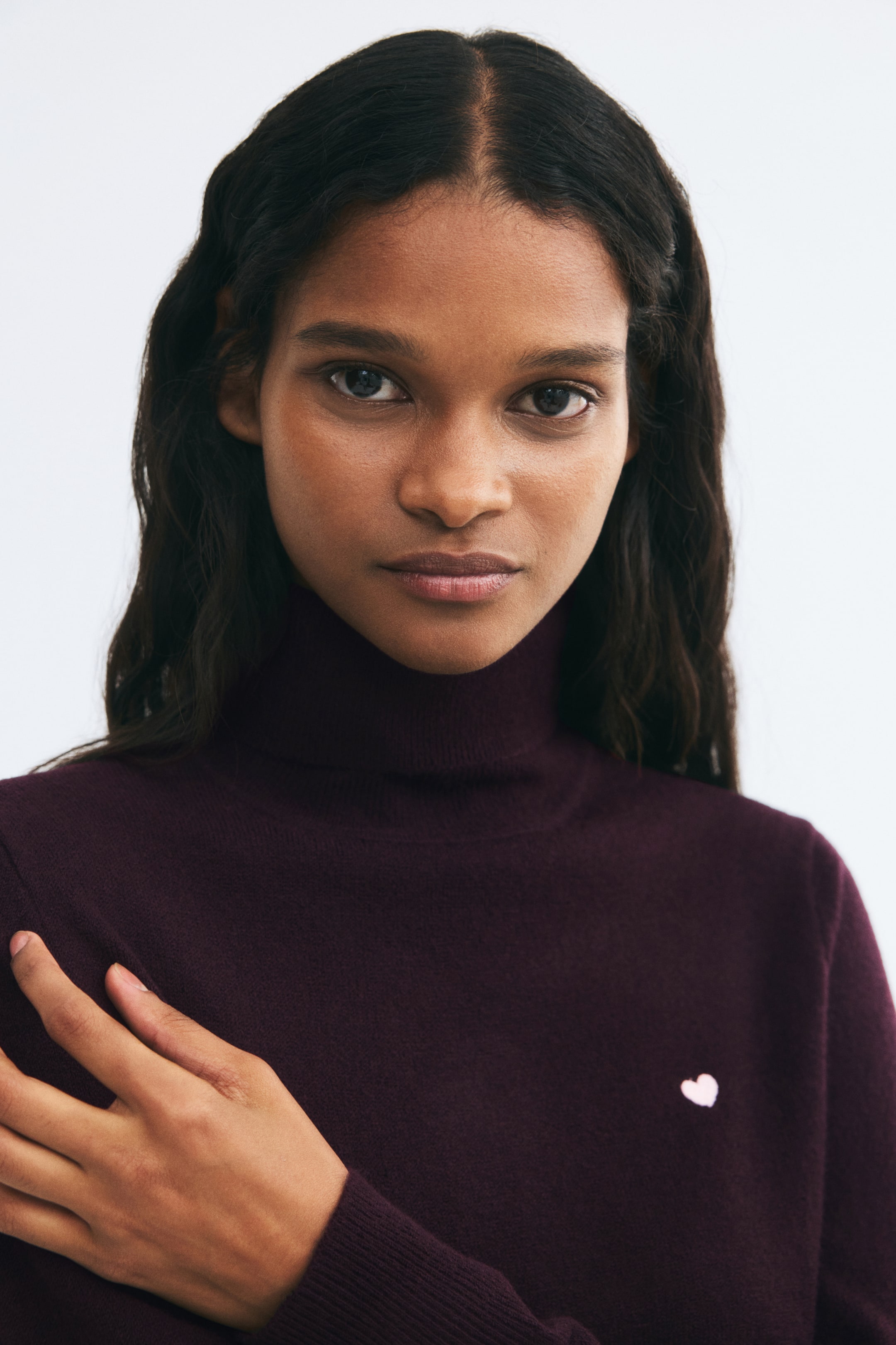 View larger image: A dark purple knitted sweater featuring a ribbed turtleneck and a small pink heart embroidered on the chest is being worn by the model. The model is resting their hand on their chest, showing the ribbed cuff of the long sleeve.