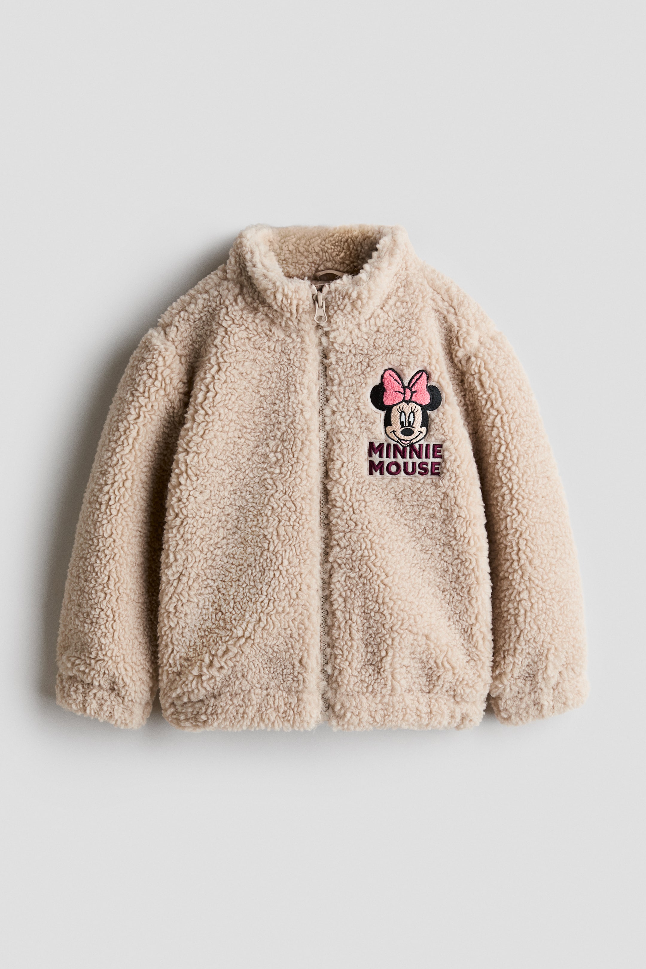 View larger image: A beige teddy fleece jacket features a textured, plush material, a full-zip front, and a stand-up collar. An embroidered Minnie Mouse patch with a pink bow and "Minnie Mouse" text is visible on the left chest, while elastic cuffs and hem shape the jacket.