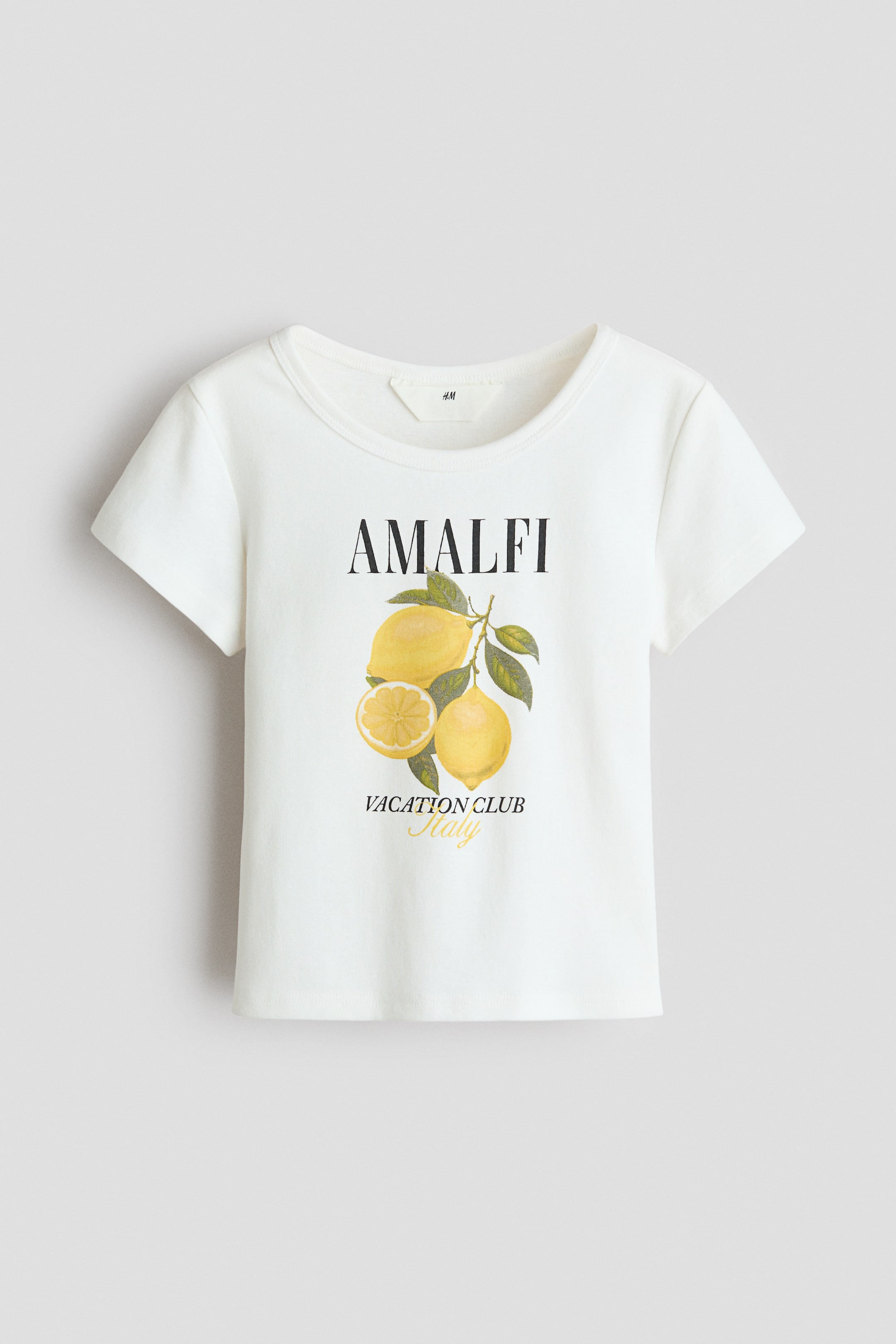 Grotere afbeelding bekijken: Een wit bedrukt jersey t-shirt met een geribde ronde halslijn wordt van voren getoond, met korte mouwen. Het toont een grafische print van de tekst "AMALFI" boven een illustratie van drie citroenen aan een tak, waarvan er één is gesneden, met daaronder "VACATION CLUB Italy".