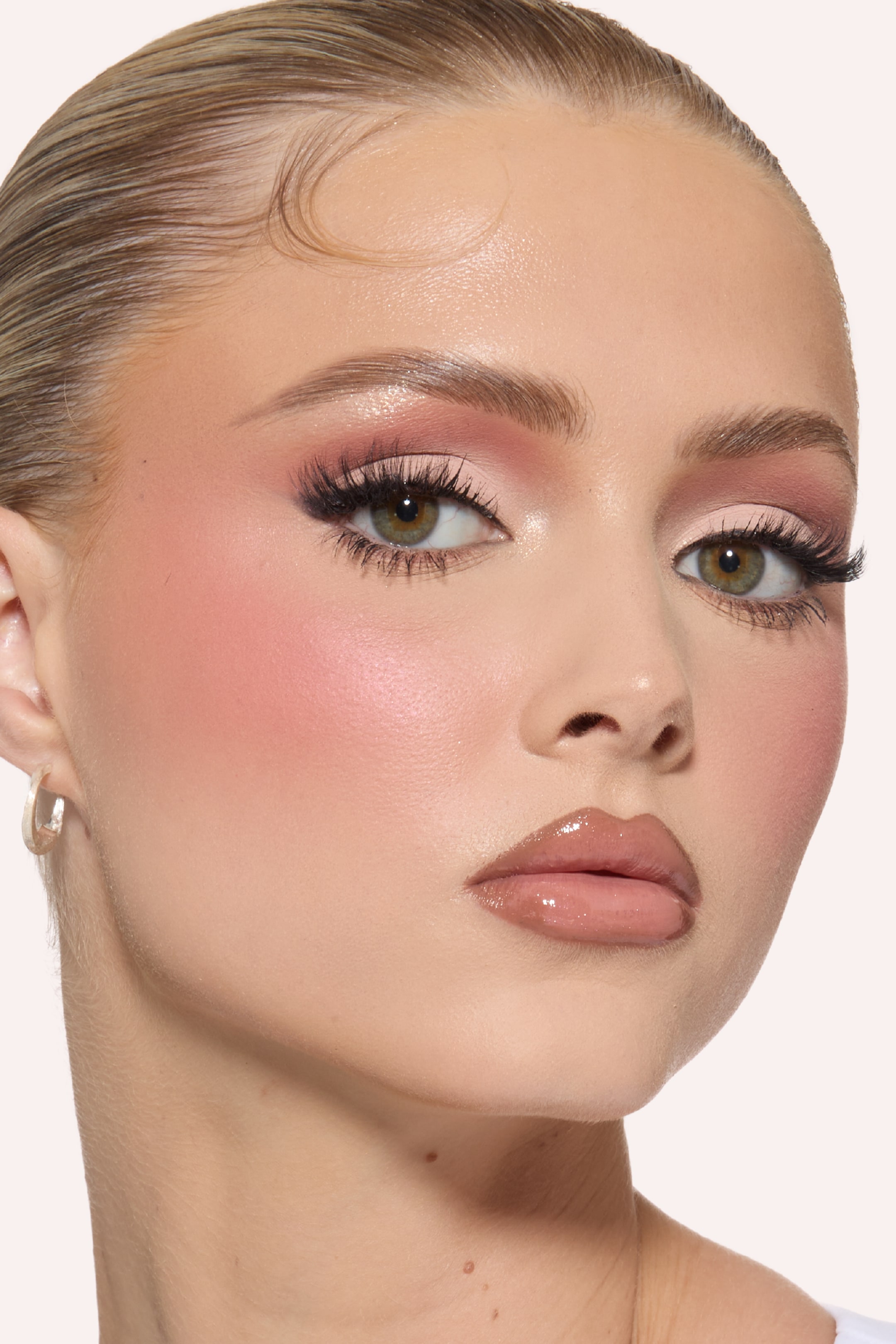 Vis større bilde: Rosa og mauve øyenskygge fremhever øynene, som også har mørk, vinget eyeliner og fyldige vipper. En rosenrød blush stryker over kinnbena, mens en blank, nude leppefarge dekker leppene. Ansiktet er lett vinklet mot høyre, og fremhever sminkens påføring.
