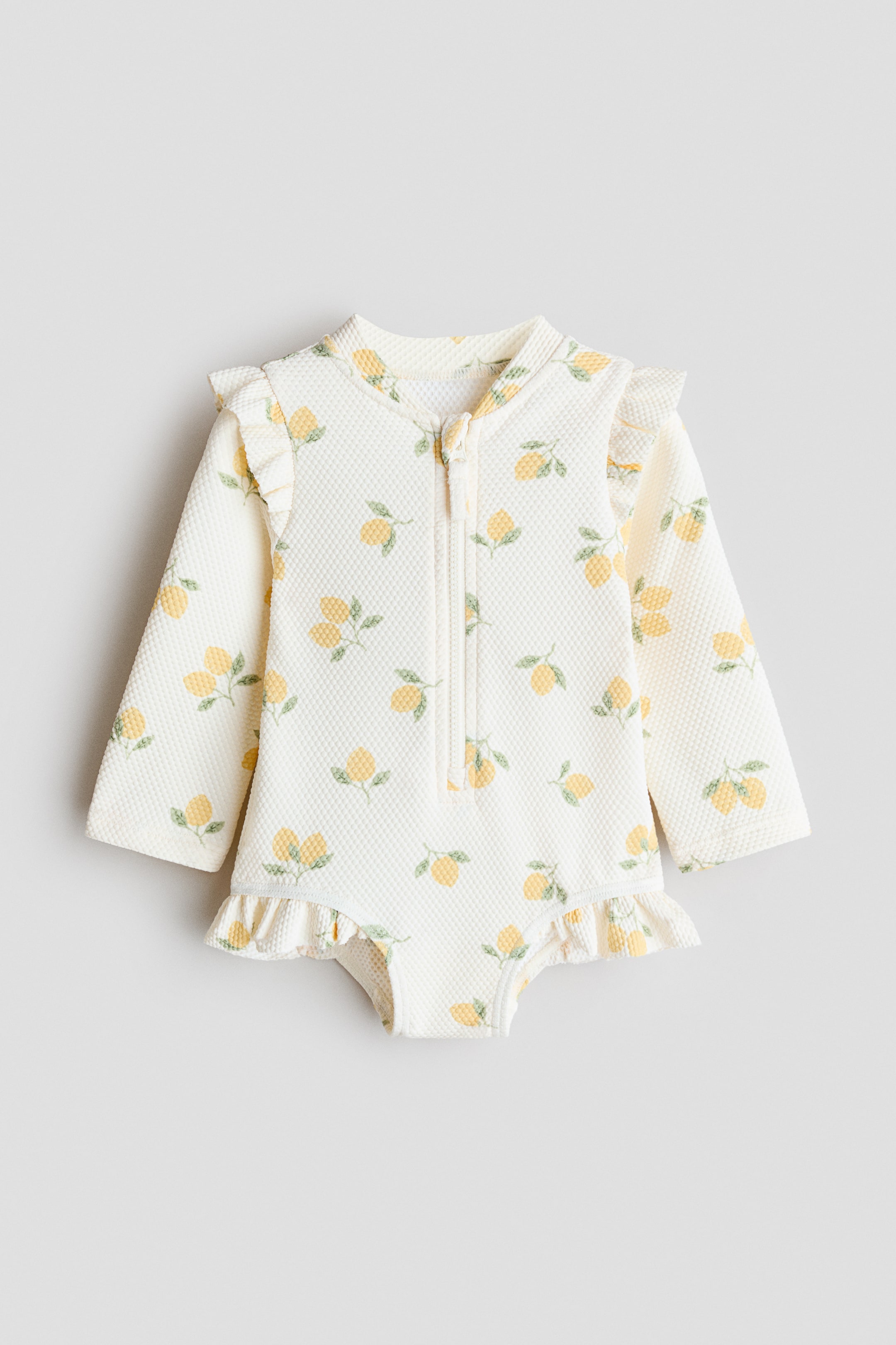 Zobacz większy obraz: Strój kąpielowy z długim rękawem, ozdobiony nadrukiem cytryn na całej powierzchni, wykonany z kremowej tkaniny o strukturze waflowej, prezentowany z przodu. Zaprojektowany z małym stójką, zamkiem błyskawicznym z przodu oraz falbankowymi detalami na ramionach i wycięciach na nogi.