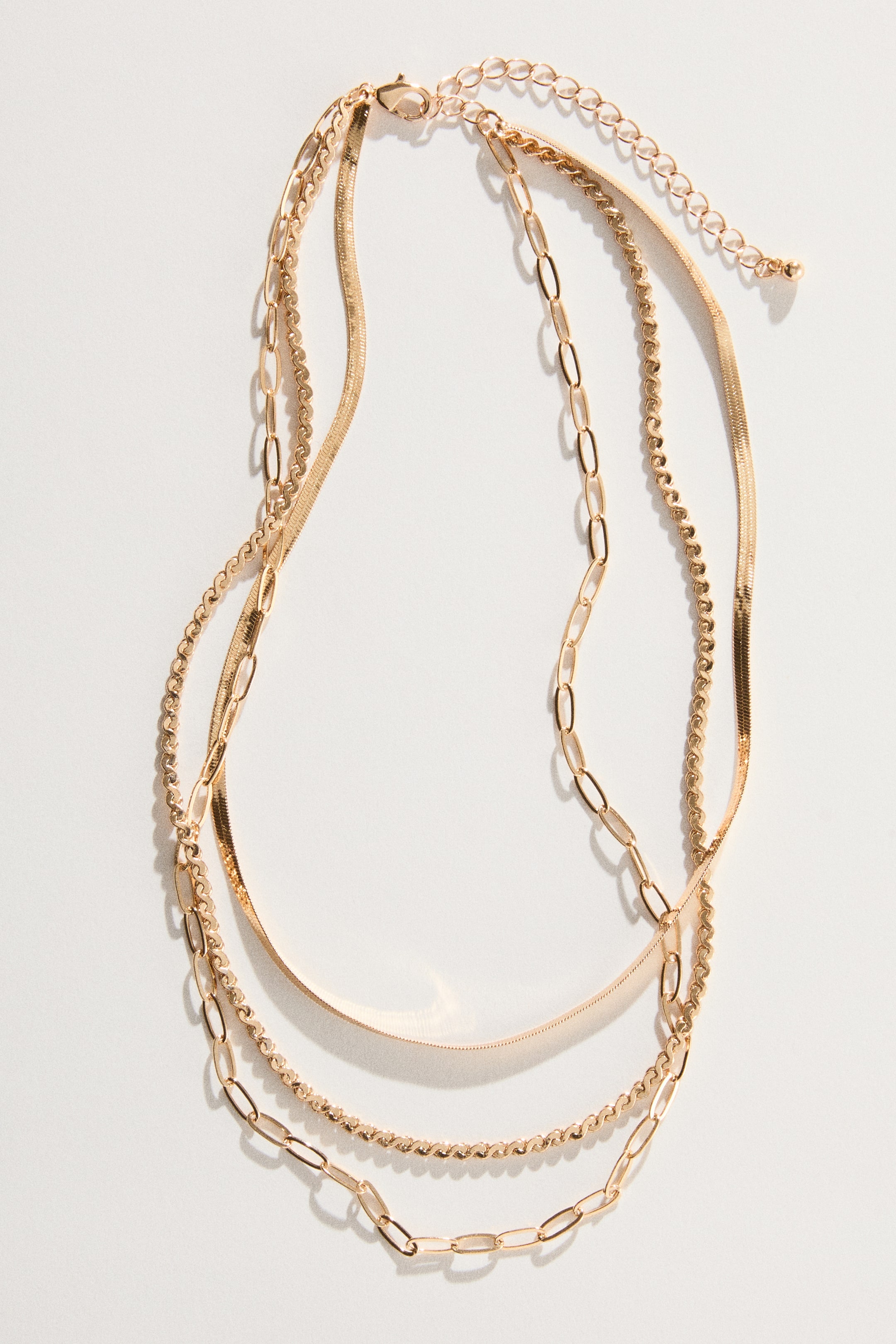 View larger image: A layered metal necklace is displayed with three chains in different designs, laid flat and facing front. It features a delicate beaded chain, a flat snake chain, and a chain with elongated oval links, all attached to an adjustable-length trigger clasp.