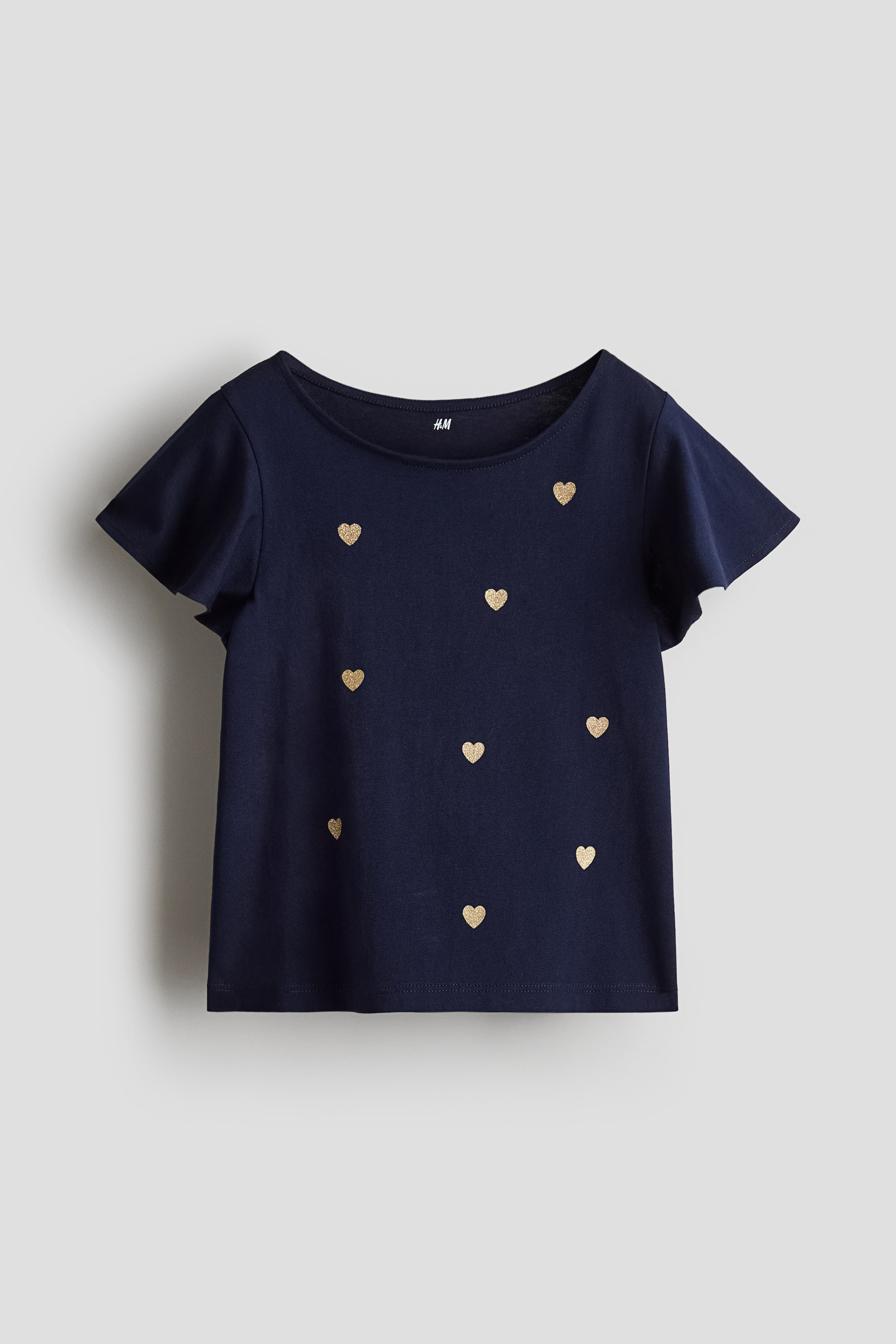 View larger image: A dark blue top in soft cotton jersey with a round neckline and flutter sleeves is displaying its front. The top is featuring scattered golden glitter heart patterns across its surface.