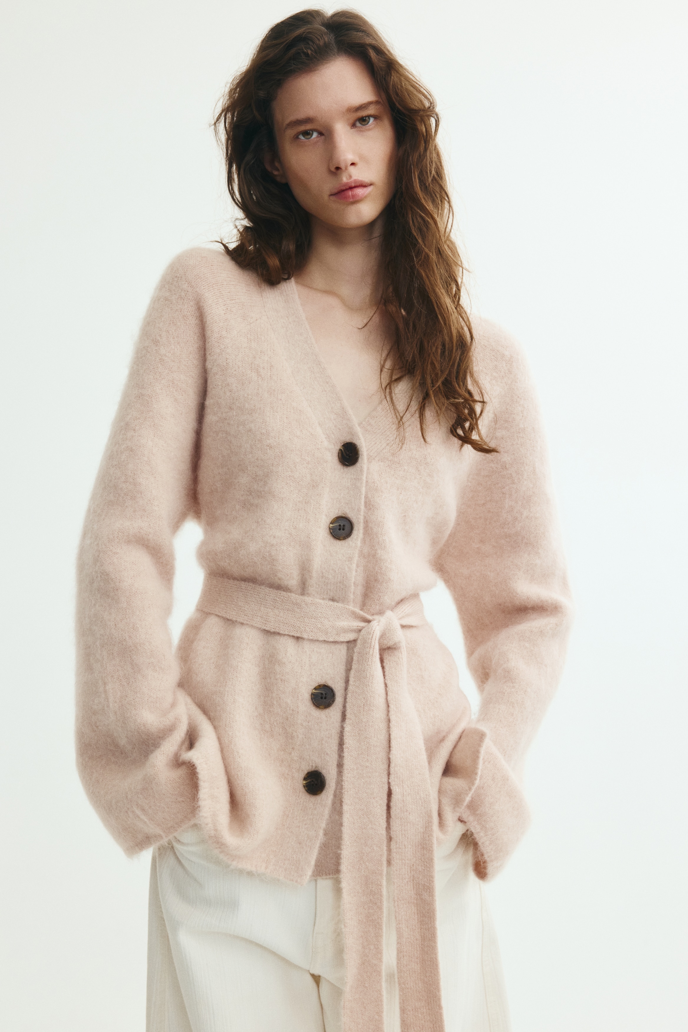 A light beige, fuzzy knitted cardigan is shown from the front, featuring a deep V-neckline, long raglan sleeves, and a front button fastening with five dark buttons. A matching knitted belt is cinched at the waist in a soft knot, accentuating the garmentâs relaxed, textured structure.