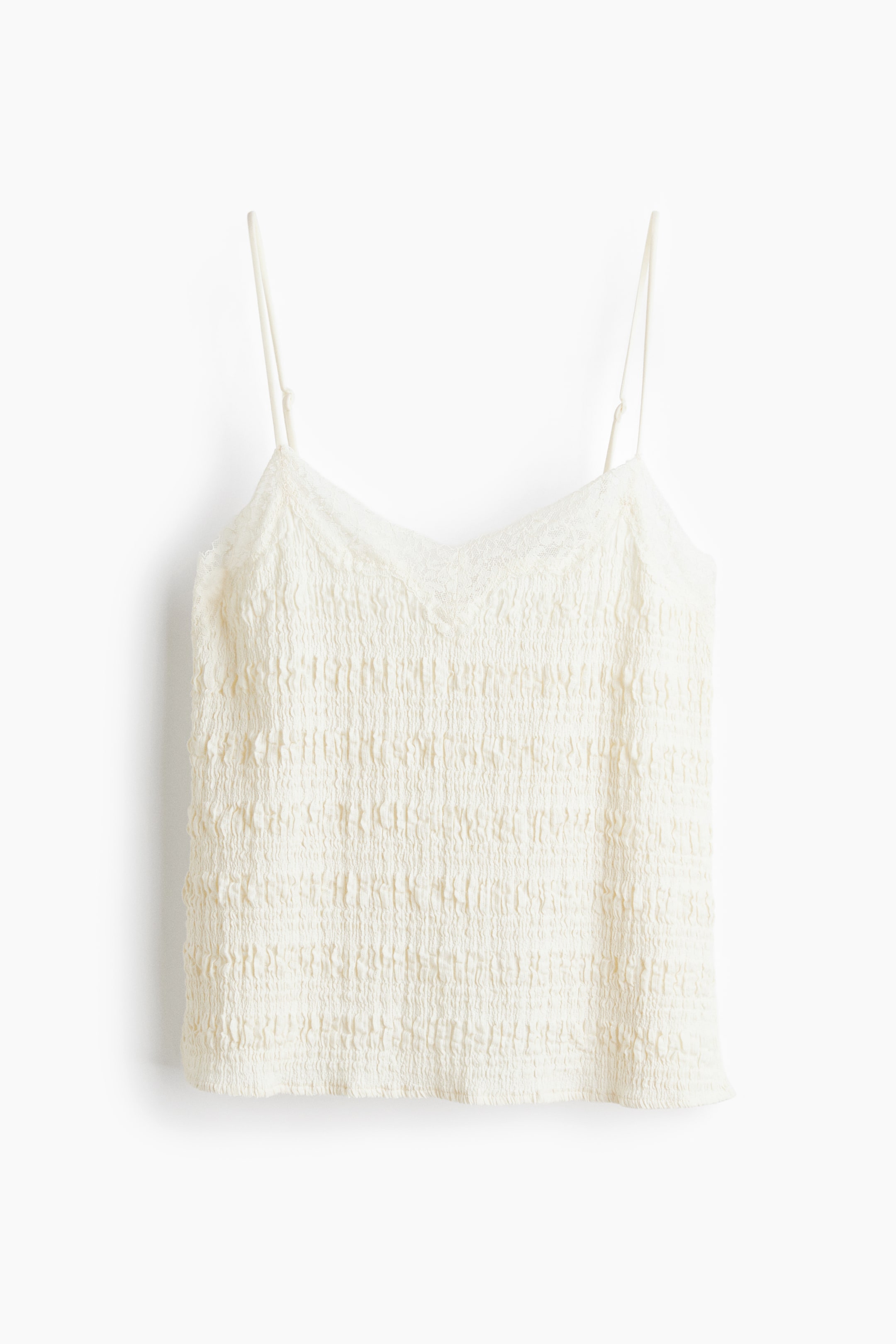 Grösseres Bild ansehen: Ein cremefarbenes Camisole-Top zeigt seine Vorderansicht, mit einem gesmokten Körper und einem V-Ausschnitt, der mit zarter Spitze verziert ist. Dünne, verstellbare Spaghettiträger sind ebenfalls sichtbar.