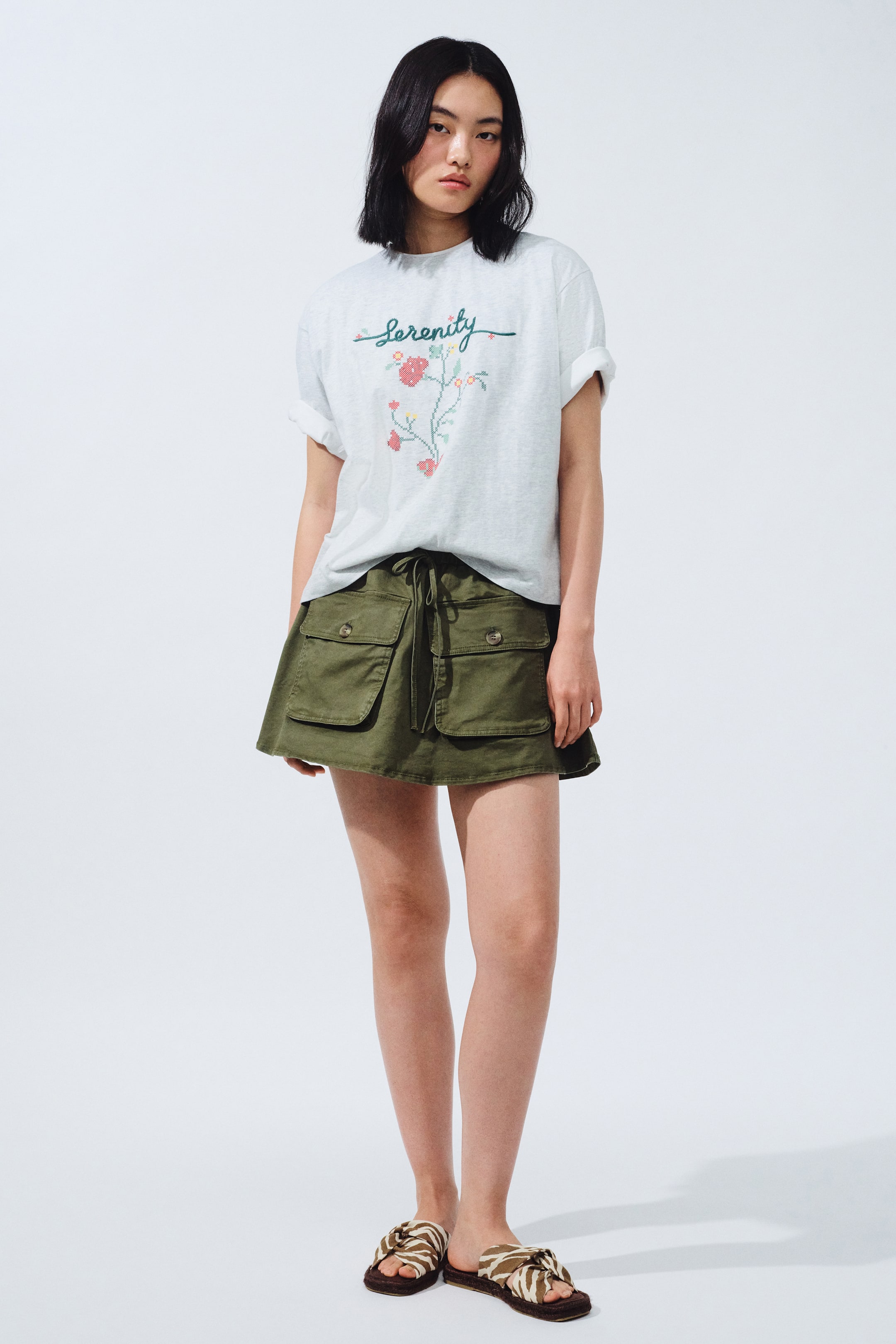 View larger image: A light grey crew neck t-shirt features short, cuffed sleeves with "Serenity" embroidery above a pixelated floral design. It is styled with an olive green A-line mini skirt, designed with a drawstring waist and two front cargo pockets with buttoned flaps. Completing the look are brown and off-white zebra-print slide sandals.