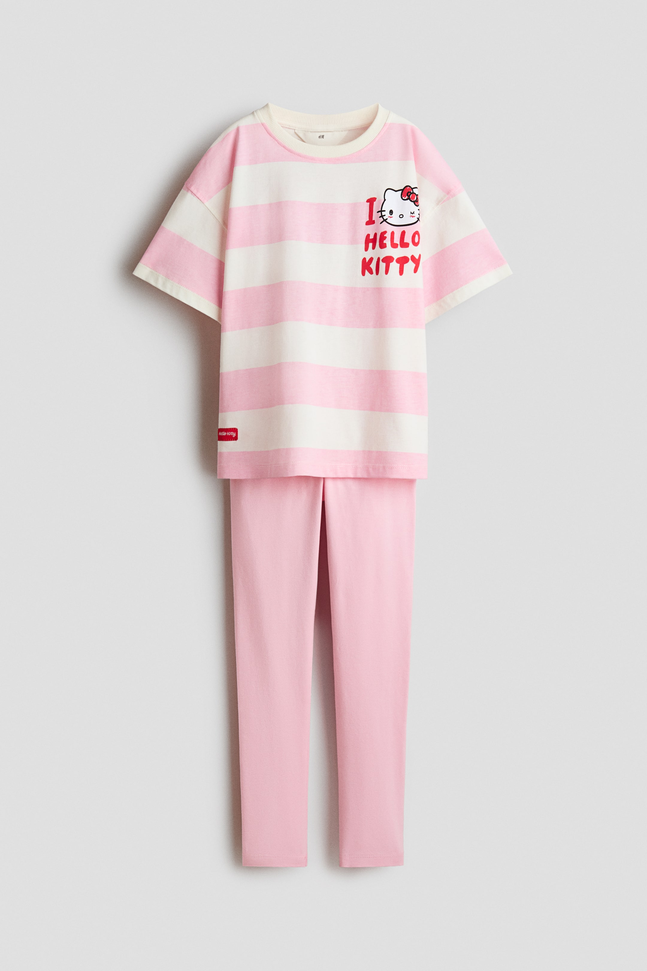 Xem ảnh lớn hơn: Một bộ cotton 2 món đang được trưng bày ở tư thế nhìn thẳng, gồm áo thun ngắn tay và quần dài. Áo thun có họa tiết sọc ngang màu hồng phấn nhạt và trắng ngà, cổ tròn gân màu kem, cùng họa tiết Hello Kitty với hình đầu nhân vật đeo nơ đỏ và dòng chữ "I ? HELLO KITTY" màu đỏ ở ngực trái, trong khi quần đi kèm có màu hồng phấn nhạt trơn.