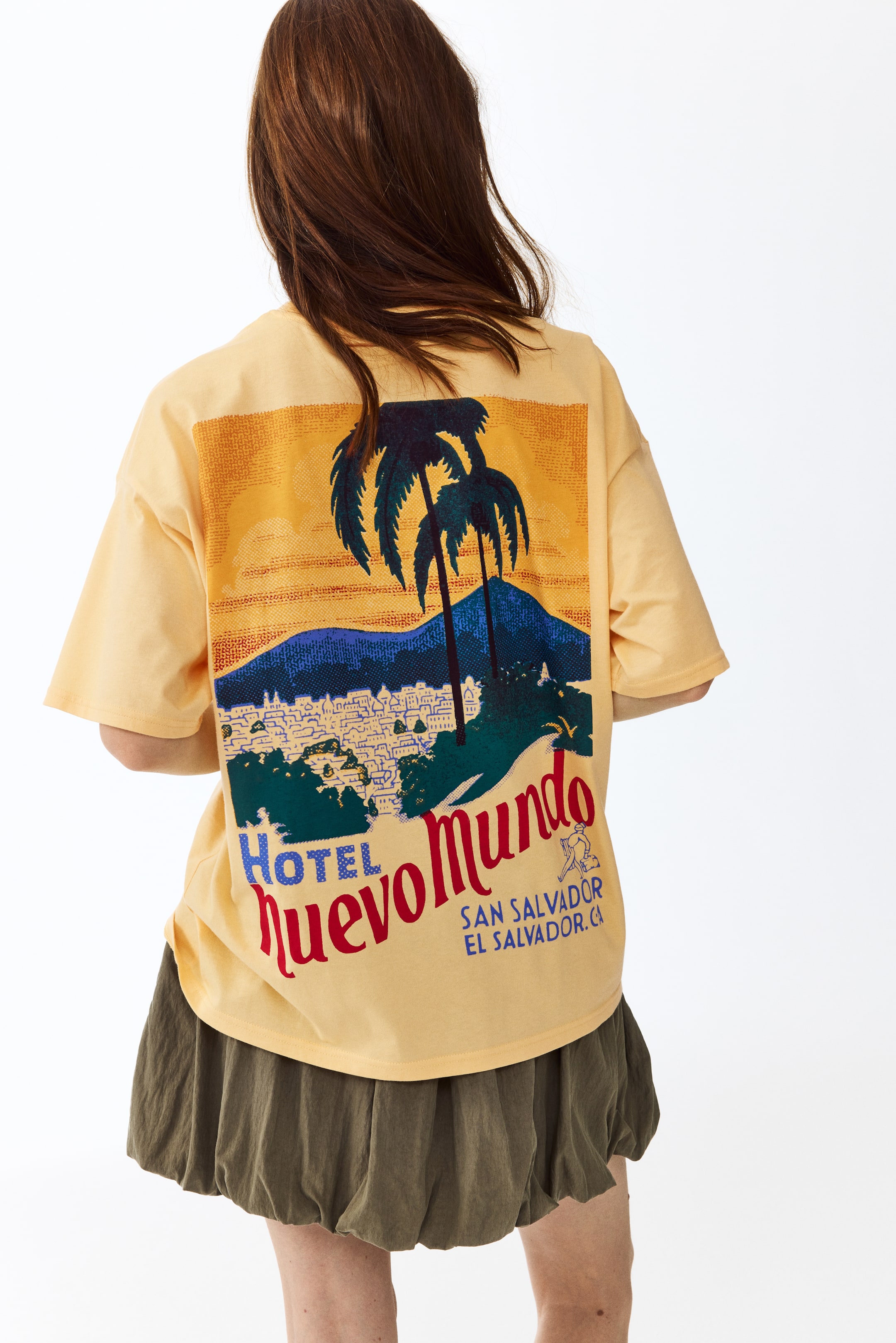 拡大画像を表示: オーバーサイズのライトオレンジTシャツが背面から示されています。大きなグラフィックプリントが特徴です。このプリントには、オレンジと黄色の空に対して2本の暗いヤシの木が描かれ、青と緑の山々と都市のスカイラインがその下に広がっています。「Hotel Nuevo Mundo」という赤と青の文字と、「San Salvador El Salvador. CA」という文字の横に小さなスーツケースを持った人物が描かれています。