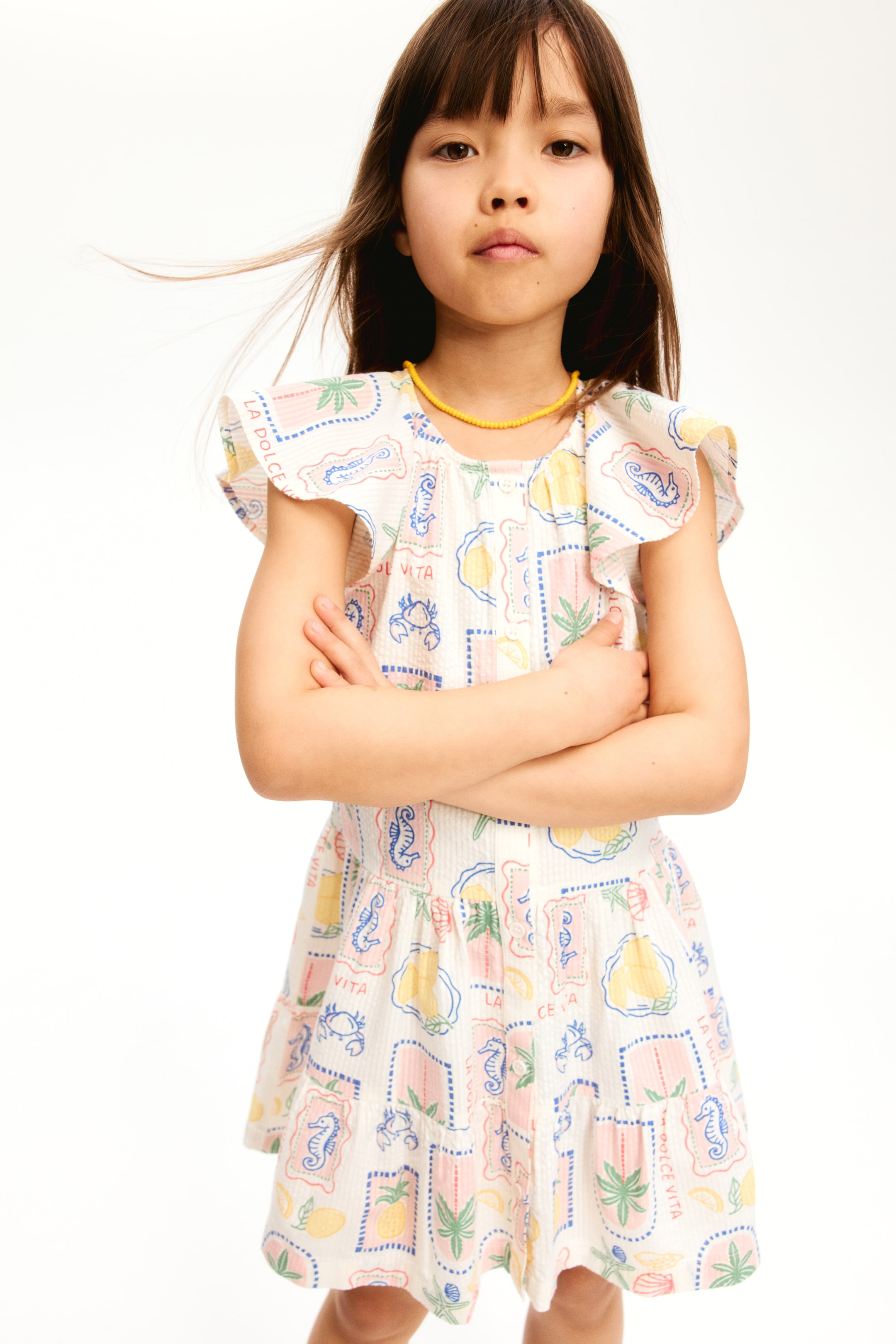 View larger image: An off-white children's short-sleeved dress showcasing a vibrant pattern of squares, each adorned with illustrations of lemons, pineapples, crabs, seahorses, and palm trees, accompanied by the text 'LA DOLCE VITA.' This textured dress is crafted with ruffled sleeves, a round neckline, buttons along the front bodice, and a skirt that flares from a gathered waist. A yellow beaded necklace elegantly rests on the neckline.