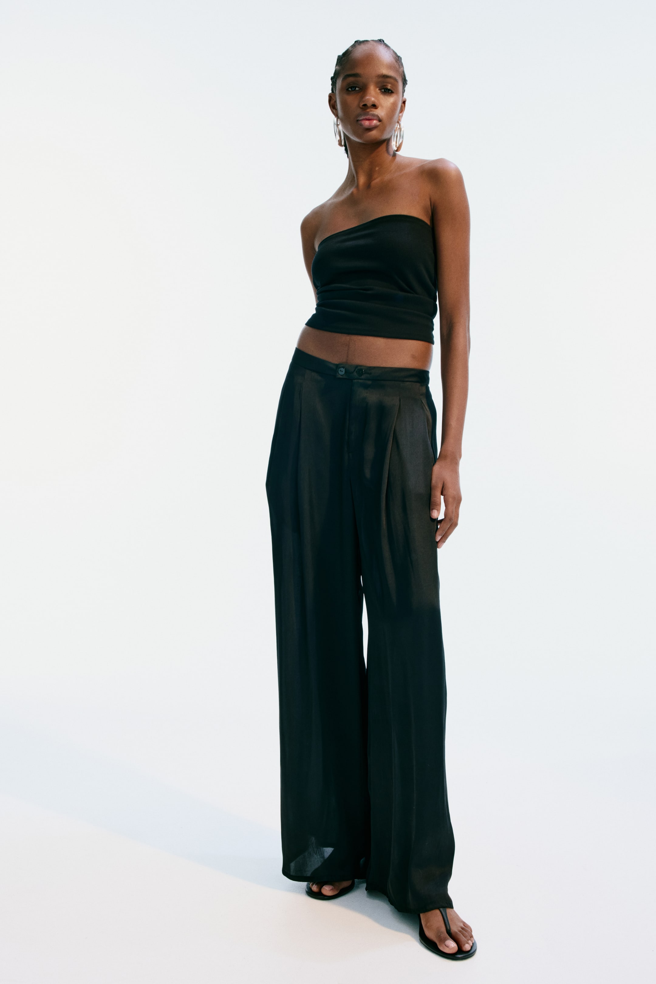 Grotere afbeelding bekijken: Losvallende broek in zachtvallend zwarte satijn wordt gedragen door het model, met een verlengde tailleband met twee zichtbare knopen, discrete zijzakken en wijde pijpen met plooien aan de bovenkant. Het model draagt de broek met een zwarte strapless top en teenslippers.
