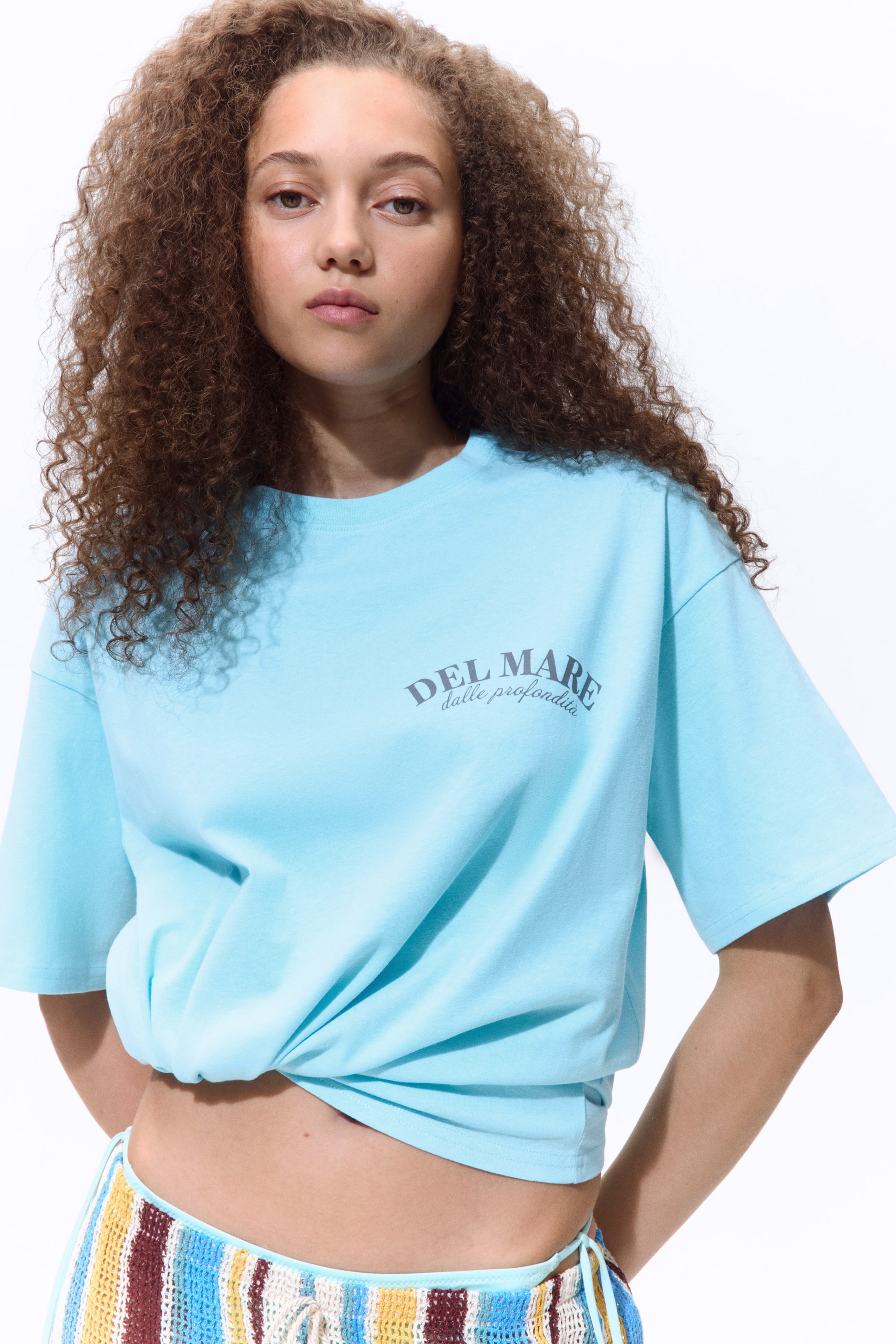 View larger image: The model is showcasing an oversized light blue soft cotton jersey t-shirt featuring a 'DEL MARE dalle profondita' print on the chest. It includes a ribbed round neckline and dropped shoulders. The t-shirt is styled with a knot at the front, revealing the midriff above a vibrant striped crochet piece.