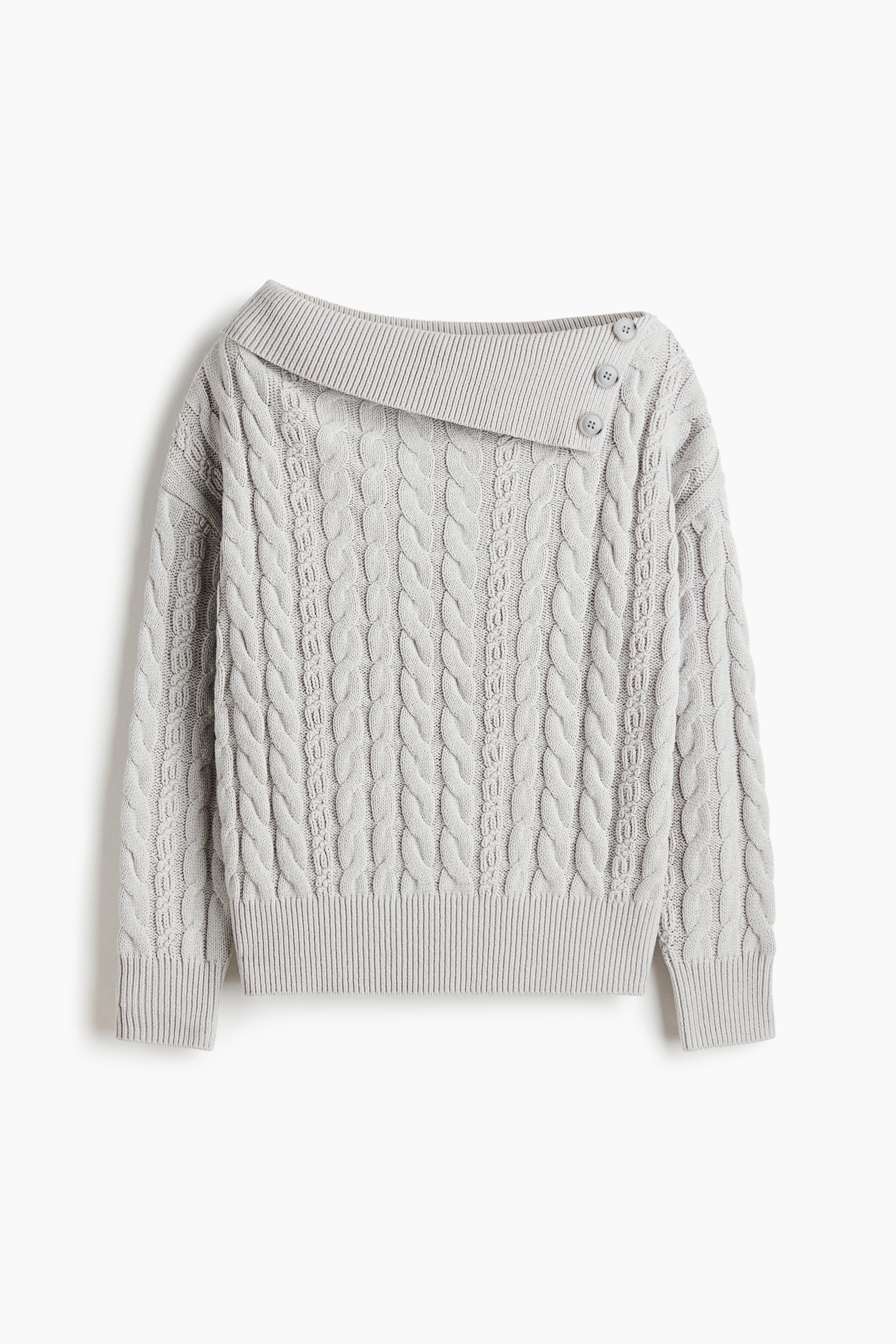 Người mẫu đang mặc một chiếc áo len dệt cáp màu xám nhạt dáng oversized, nổi bật với phần cổ gập lệch bất đối xứng có gân và ba khuy trang trí trên một bên vai. Áo được phối sơ vin vào quần đen có thắt lưng, làm nổi bật phom dáng thoải mái cùng phần bo tay áo có gân.