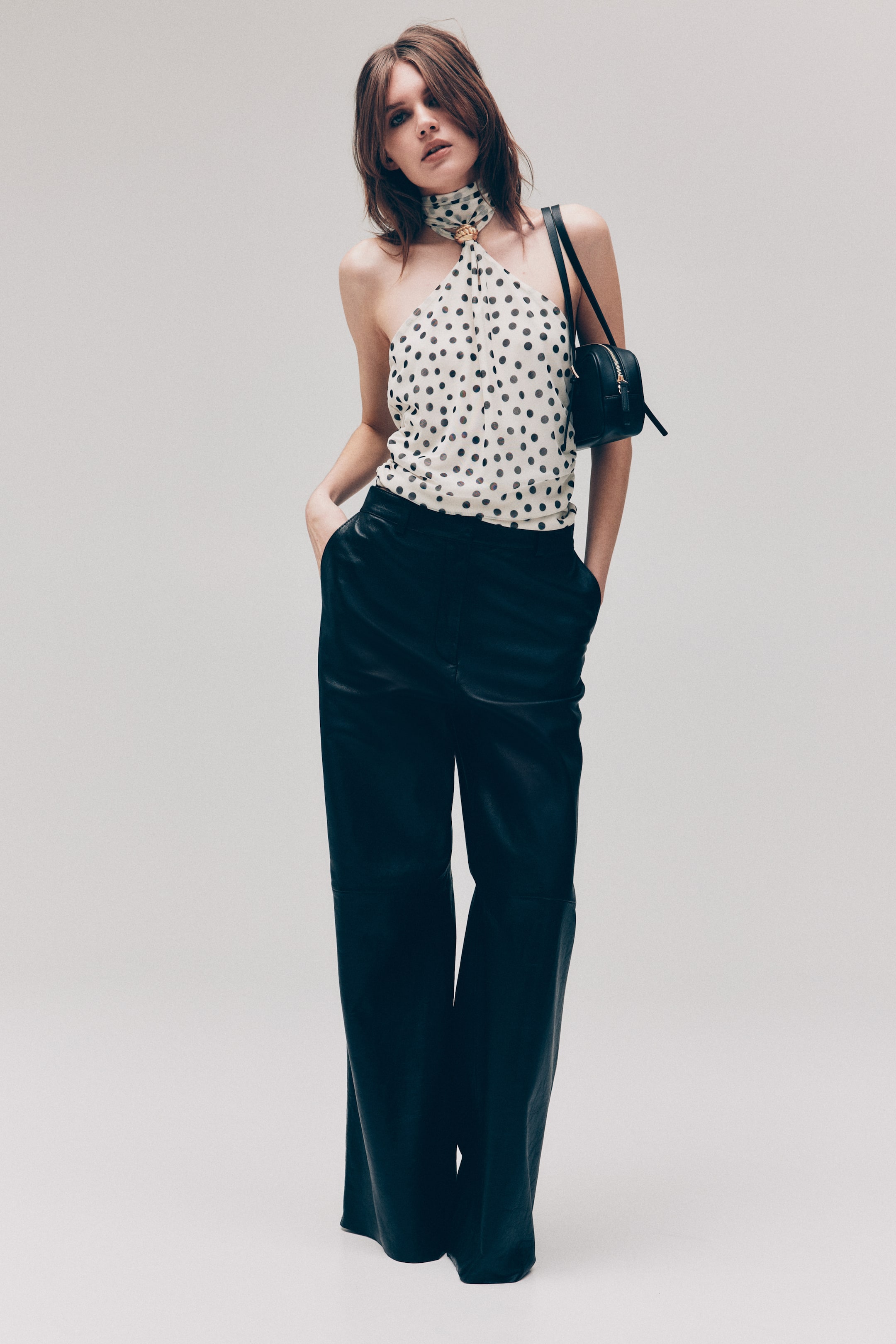 View larger image: The model is wearing a white halter top with black polka dots and a gold-tone embellishment at the neckline. She pairs it with black wide-leg trousers, with her hands resting in the pockets, and carries a small black shoulder bag over her shoulder.