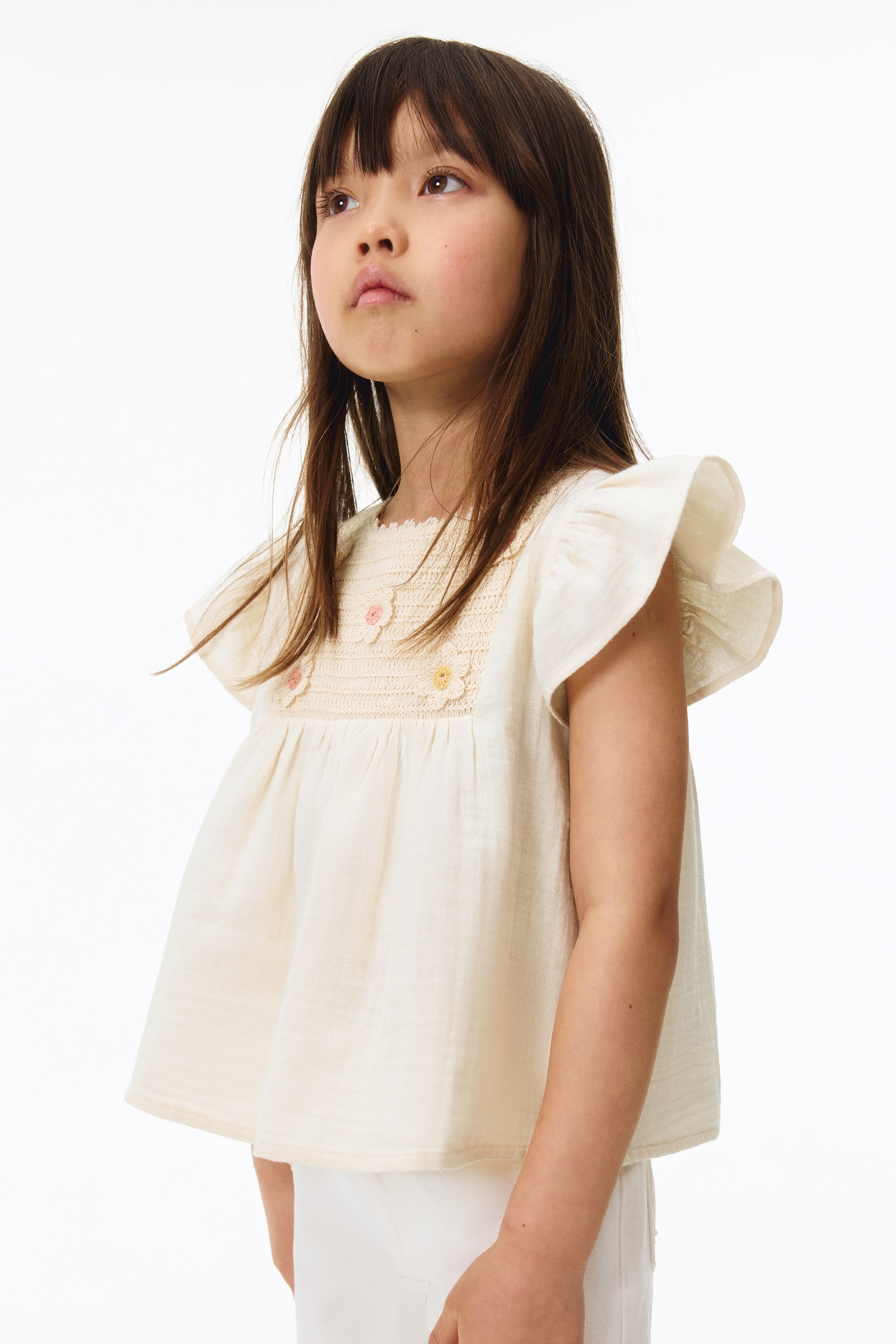 View larger image: The off-white top showcases a crocheted yoke across the chest, embellished with three small embroidered flowers. It features short, ruffled sleeves and a gathered, flowing body, offering a loose fit when viewed from a slight front-right angle.