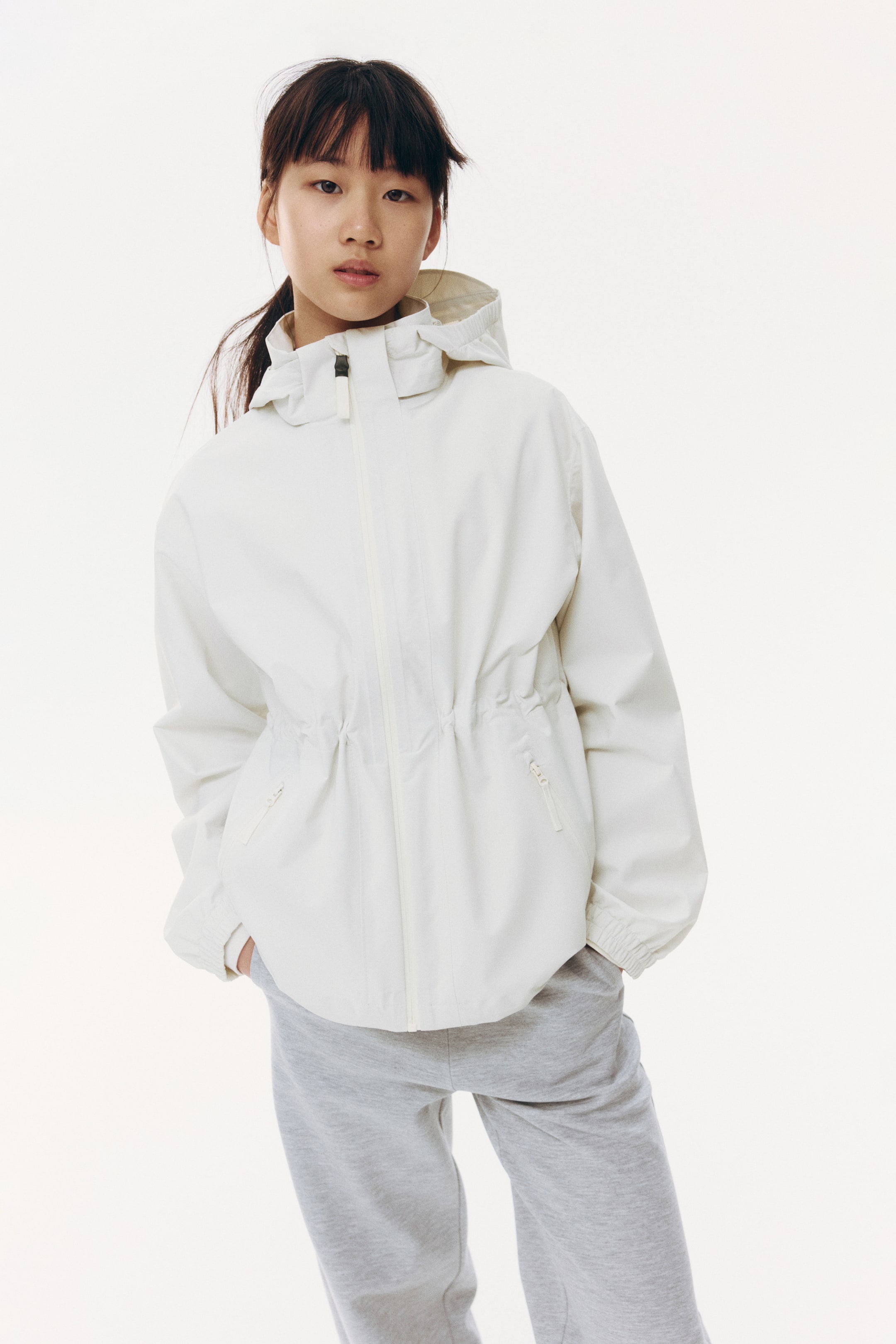 Grotere afbeelding bekijken: Een witte jas met capuchon wordt gepresenteerd met de voorkant naar voren, voorzien van een ritssluiting over de volledige lengte, een capuchon met trekkoord, een taille met trekkoord, twee voorzakken met rits en elastische manchetten.