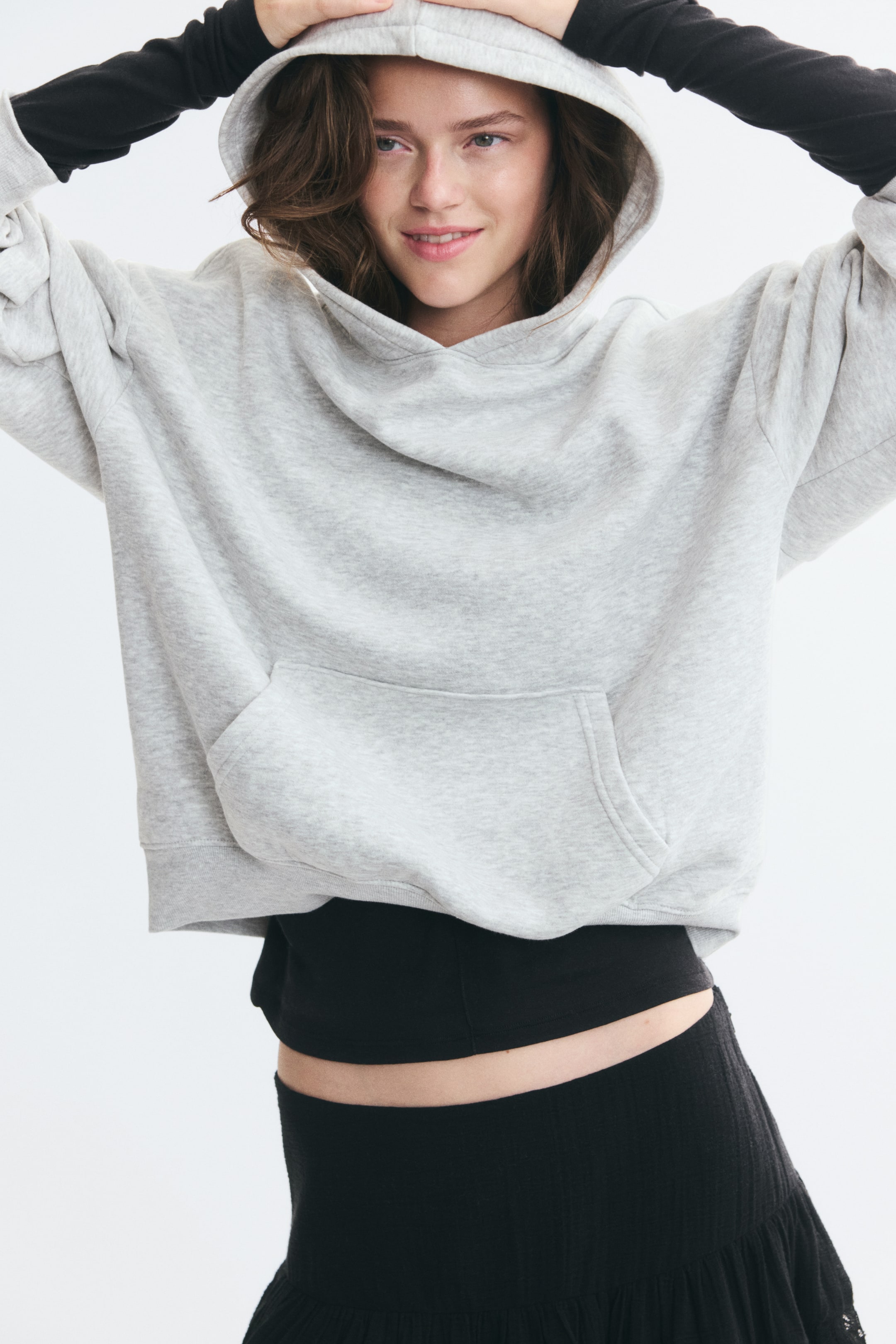 View larger image: The model is wearing a light grey fleece hoodie, holding the hood over their head with both hands. The hoodie features dropped shoulders, a kangaroo pocket and a ribbed hem, and is layered over a black long-sleeved top and black skirt.