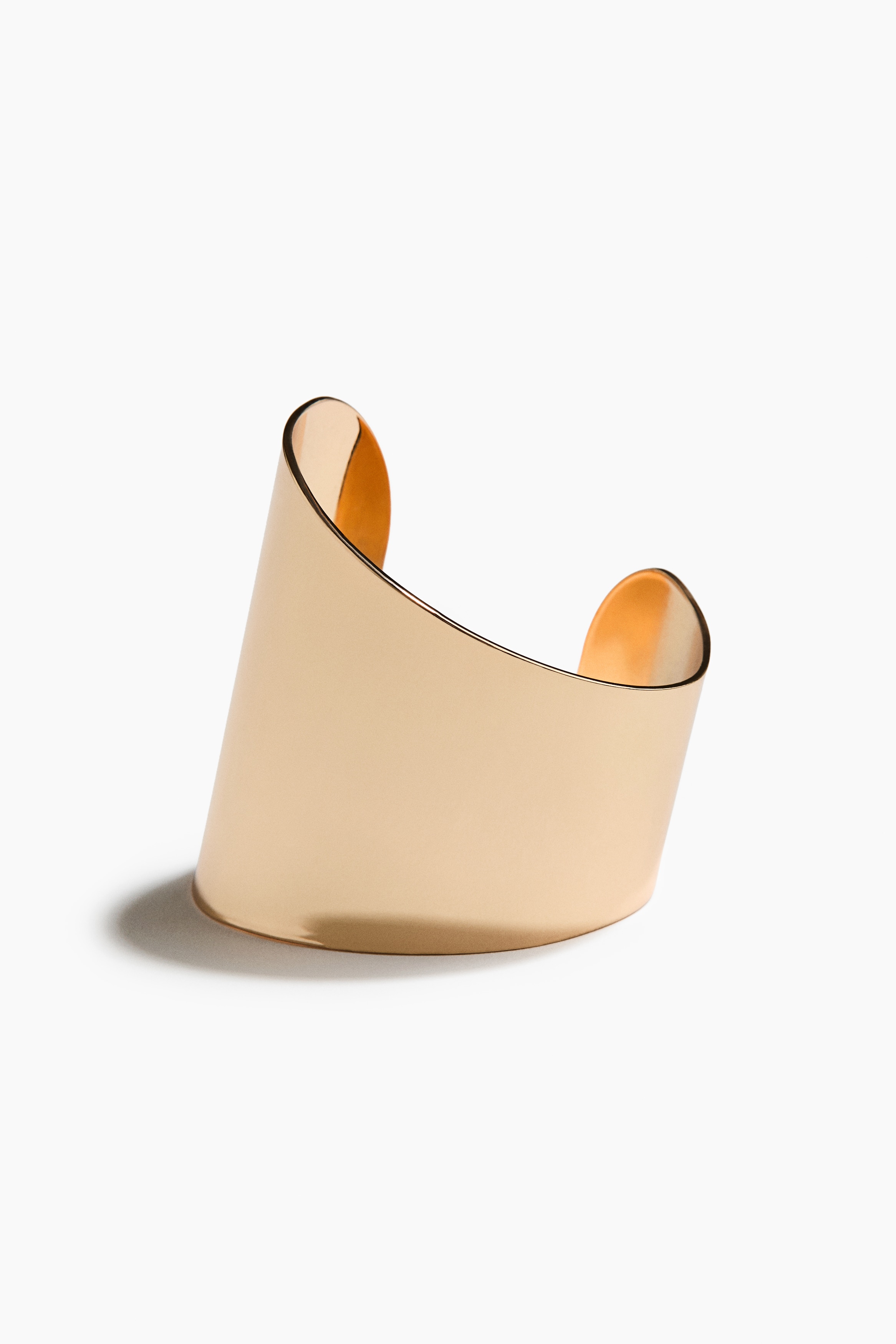 A wide, shiny gold-toned cuff bracelet with an asymmetrical design is worn on the modelâs forearm. The model rests their hand under their chin, highlighting the polished, reflective finish of the jewellery.