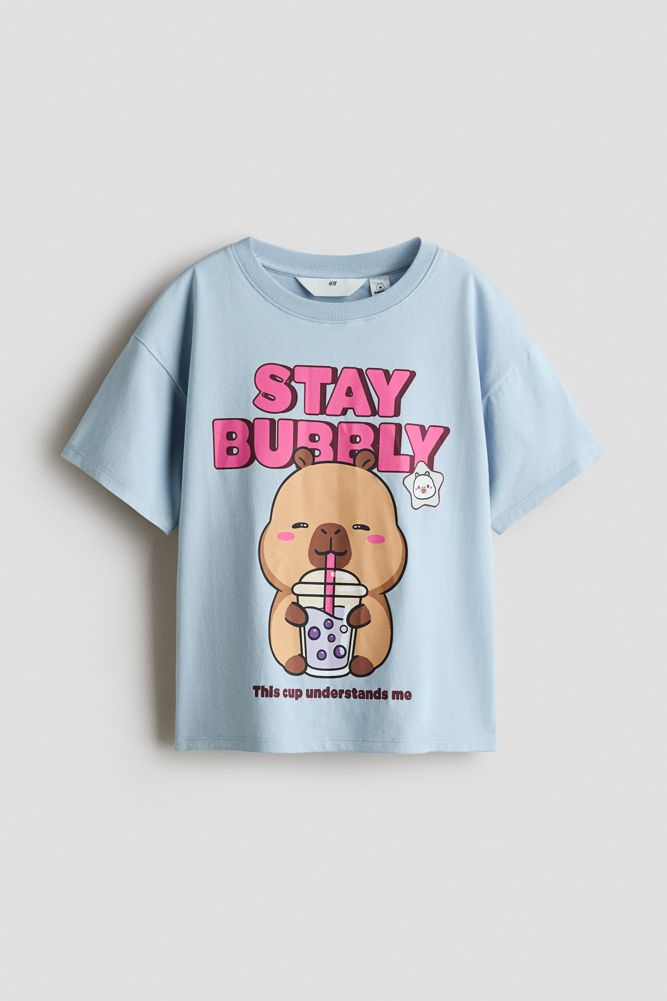 查看大圖: 一件淺藍色棉質 jersey T 恤正面展示,採用羅紋圓領與自然落肩設計。正面印有一幅可愛卡通圖案,描繪一隻水豚正在喝珍珠奶茶,圖案位於醒目的粉紅色文字「STAY BUBBLY」與較小的棕色文字「This cup understands me」之間。