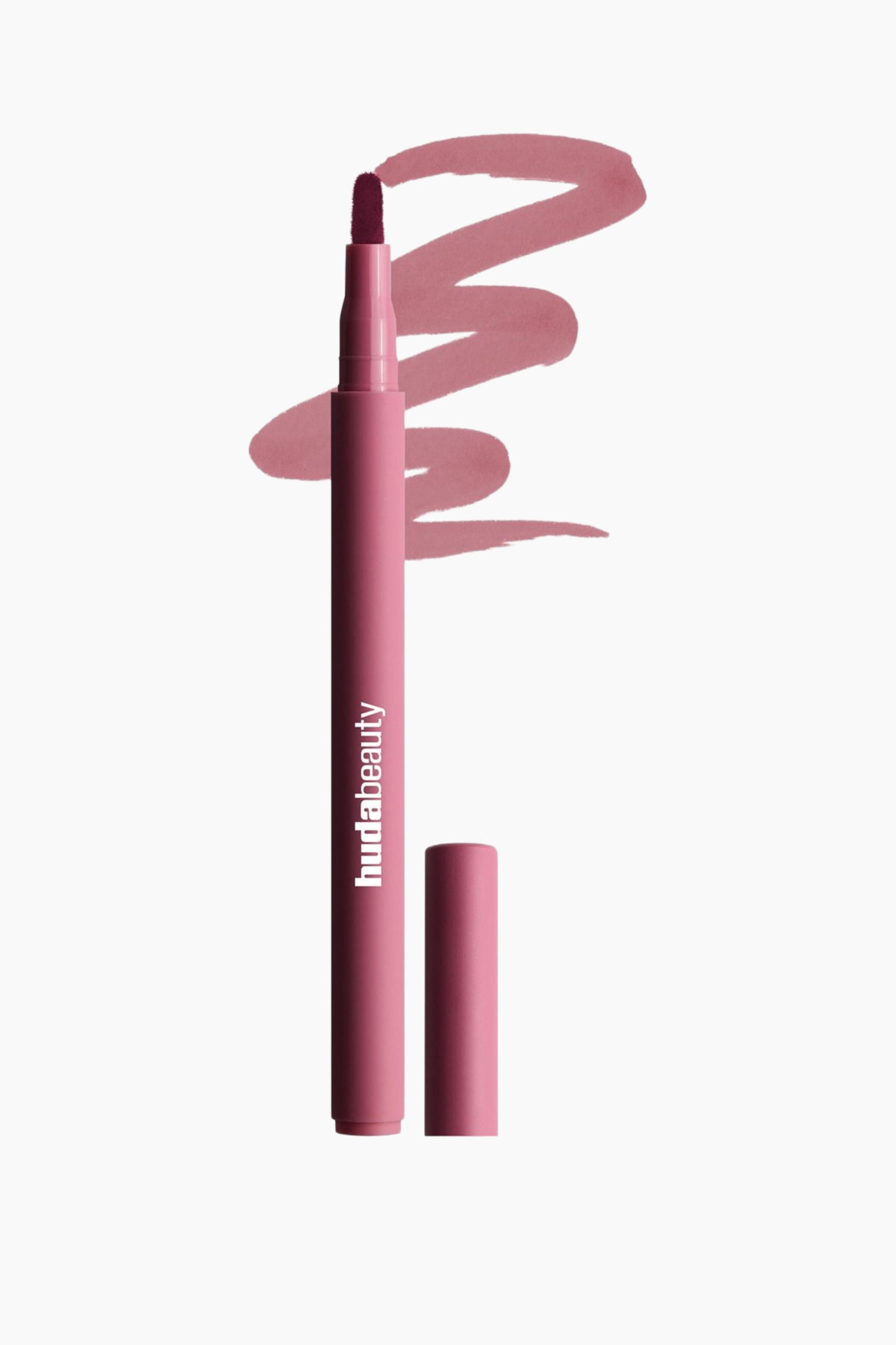 Visa större bild: En rosa flytande läppprodukt i pennformat står upprätt, lätt vinklad åt höger, med korken av och en mörkt bärfärgad filtspets synlig. Varumärket "hudabeauty" är tryckt vertikalt på sidan, och ett virvlat prov av den rosa produkten syns bakom pennan.