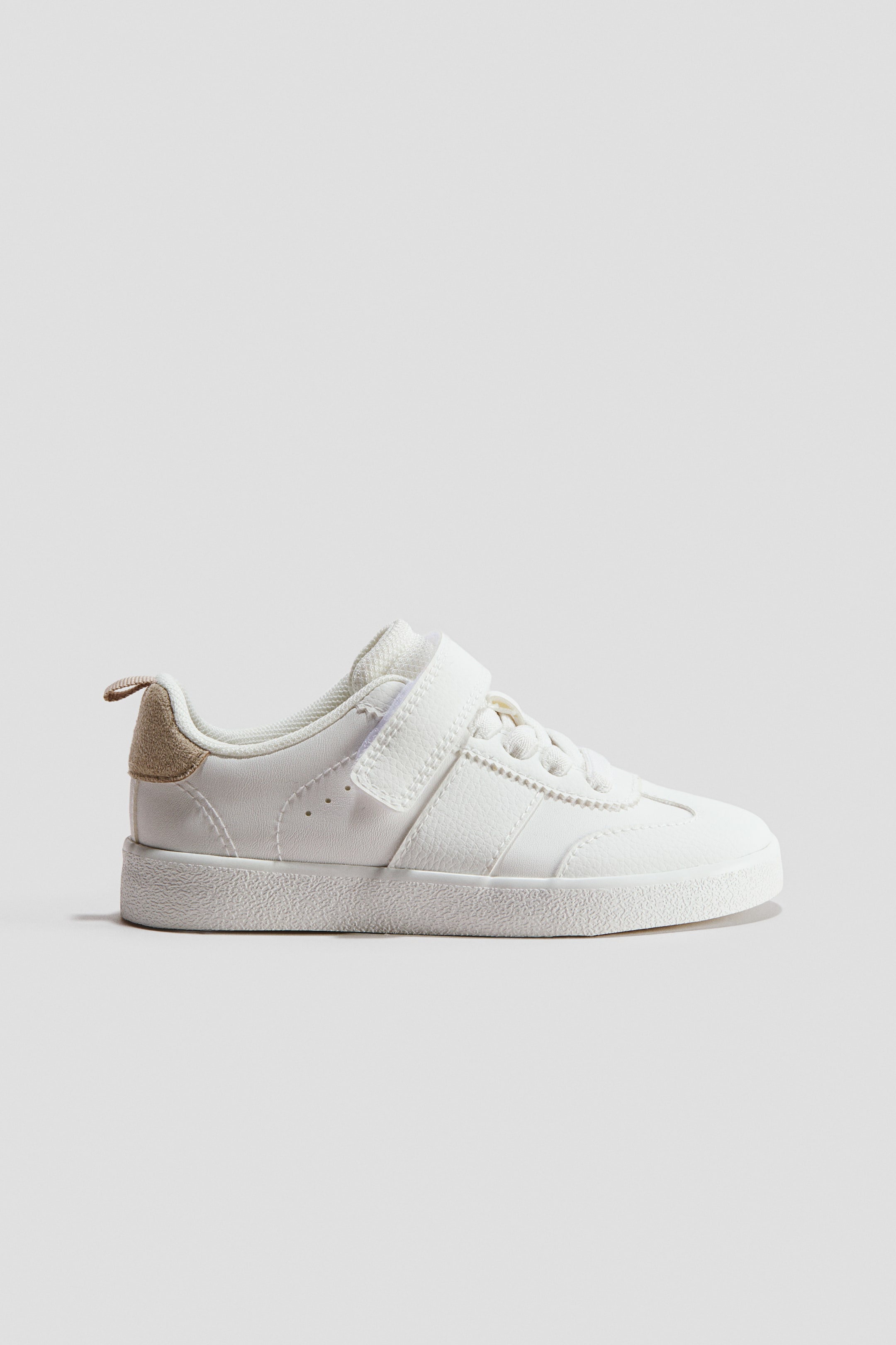 View larger image: A white sneaker is shown in side profile, featuring a single white Velcro strap over fixed white laces and eyelets. It is designed with a textured white sole and a contrasting tan heel tab.