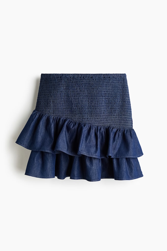 Le devant d'une jupe courte en denim bleu foncé à smocks est doté d'une large ceinture smockée et de deux volants horizontaux superposés au-dessus de l'ourlet. - 1