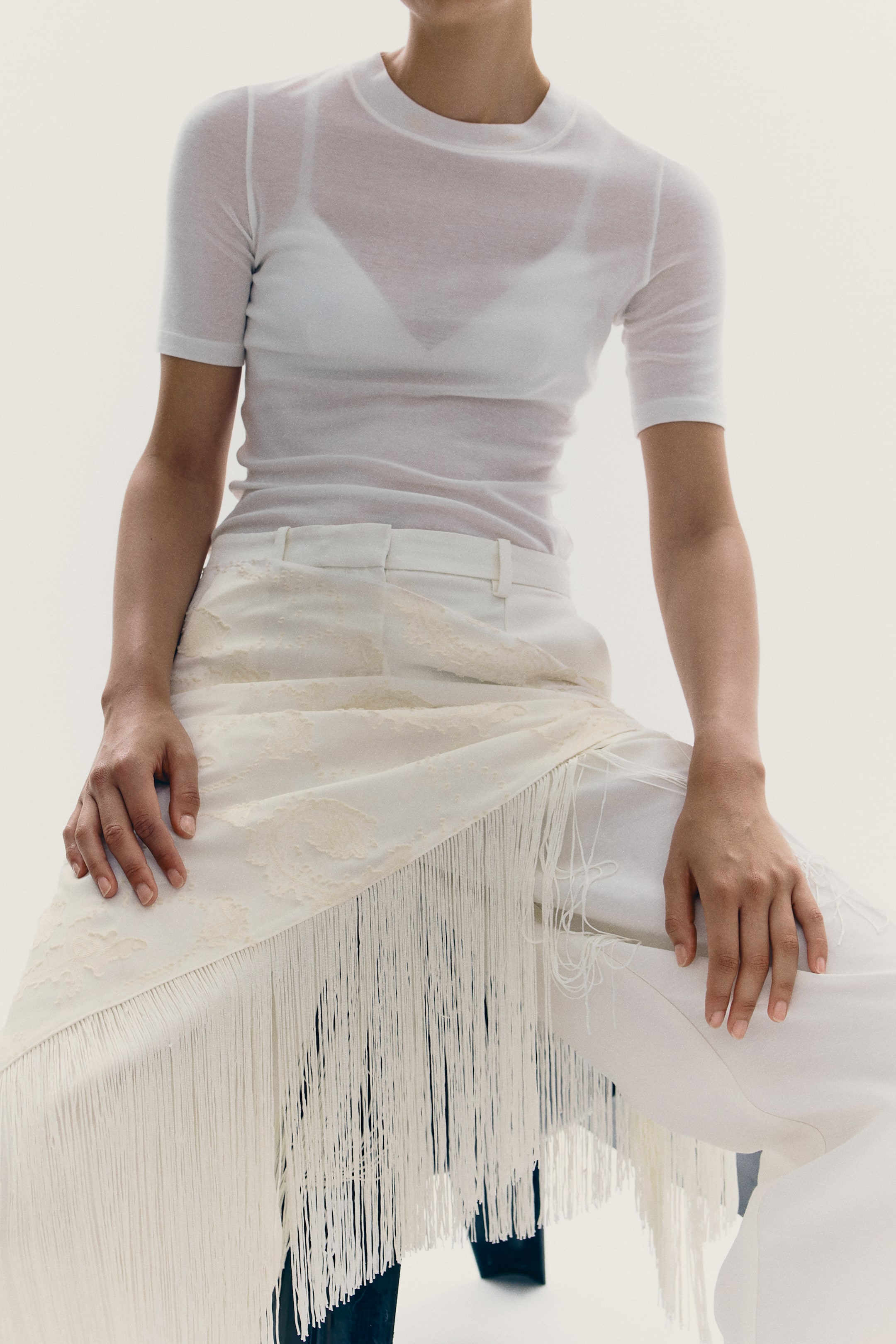 View larger image: A triangle scarf in a jacquard weave, detailed with long fringes along two edges, is draped across the model's lap. The model is resting their hands on the scarf, which is layered over white trousers.