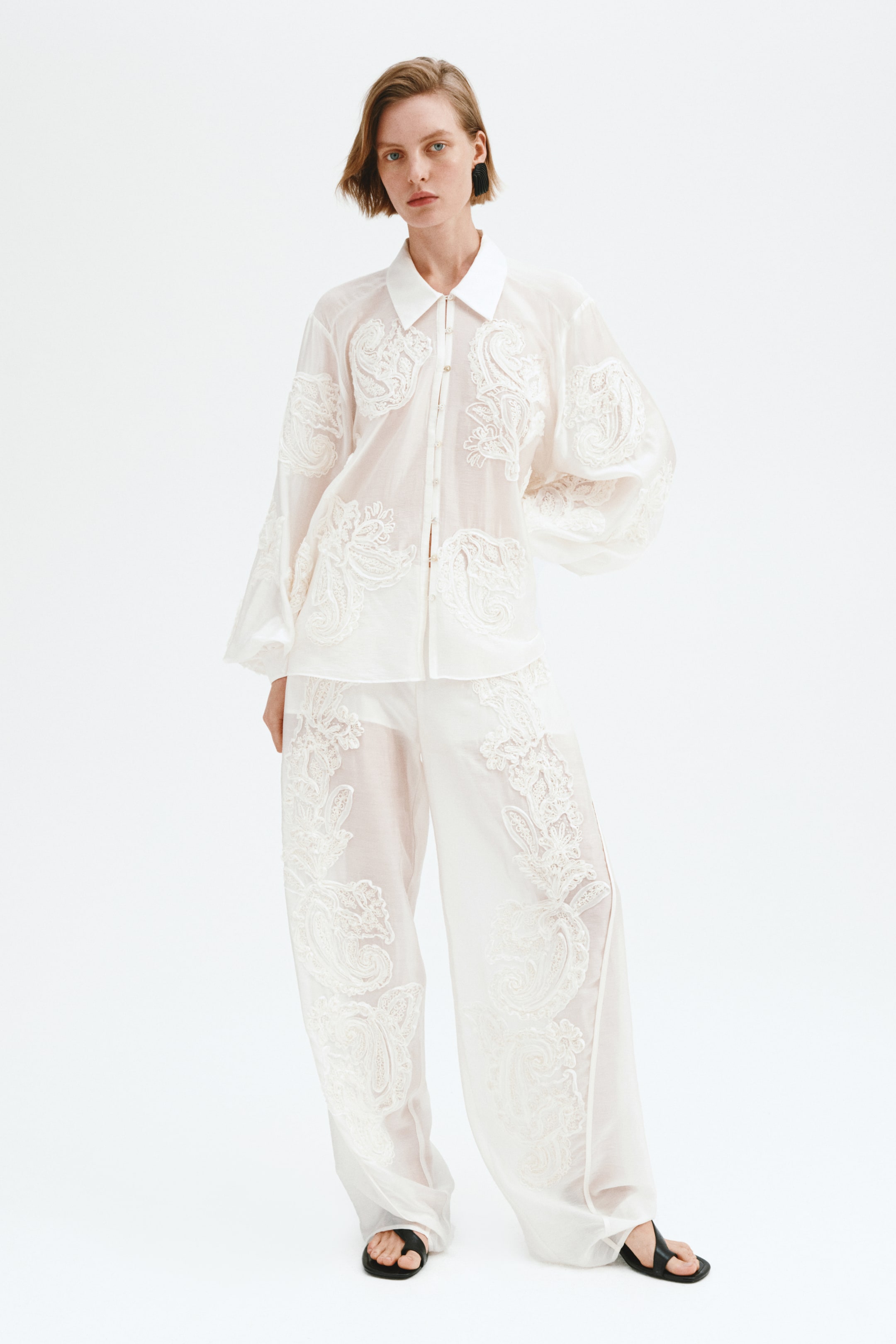 Ingrandisci l'immagine: Una camicia bianca trasparente con colletto classico e maniche lunghe arricciate viene presentata da un'angolazione frontale, caratterizzata da intricati motivi paisley ricamati in bianco. È abbinata a pantaloni trasparenti a gamba larga, anch'essi adornati con ricami simili.