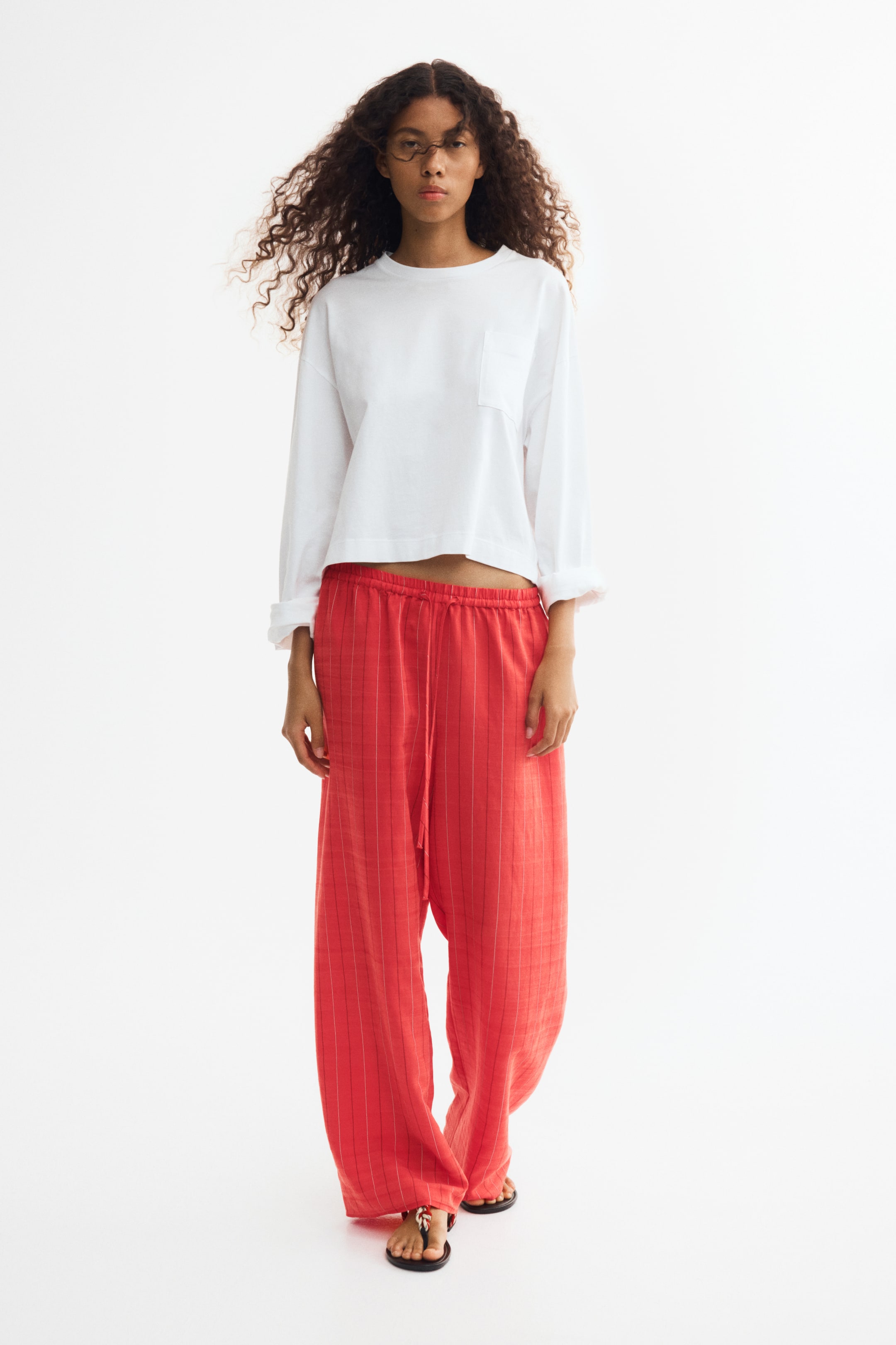 Grotere afbeelding bekijken: De model draagt een rode broek met fijne verticale strepen, een elastische tailleband met trekkoord en wijde pijpen. De lichtgewicht broek is gestyled met een witte top met lange mouwen.