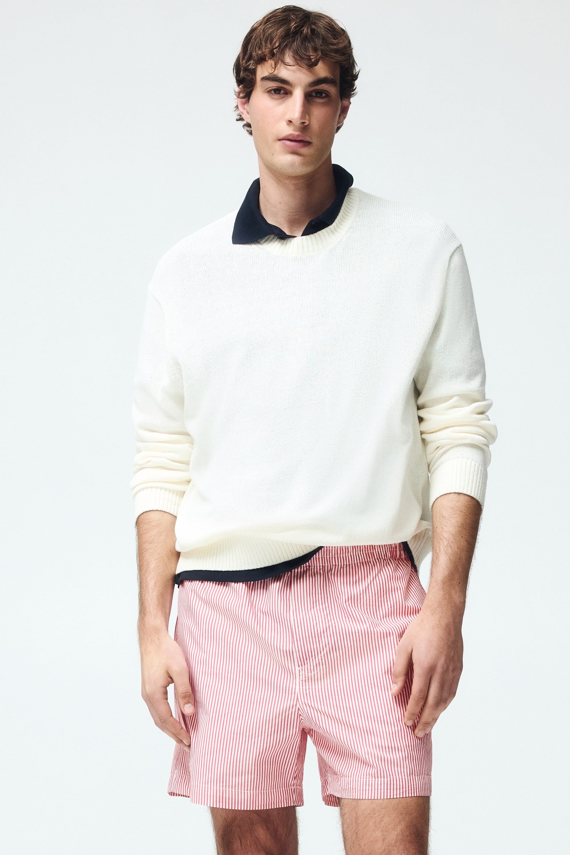 Ampliar la imagen: Un suéter blanco de cuello redondo con puños y dobladillo acanalados se está combinando sobre una camisa polo azul marino con el cuello visible, y unos shorts a rayas verticales rojas y blancas con cintura elástica completan el conjunto.