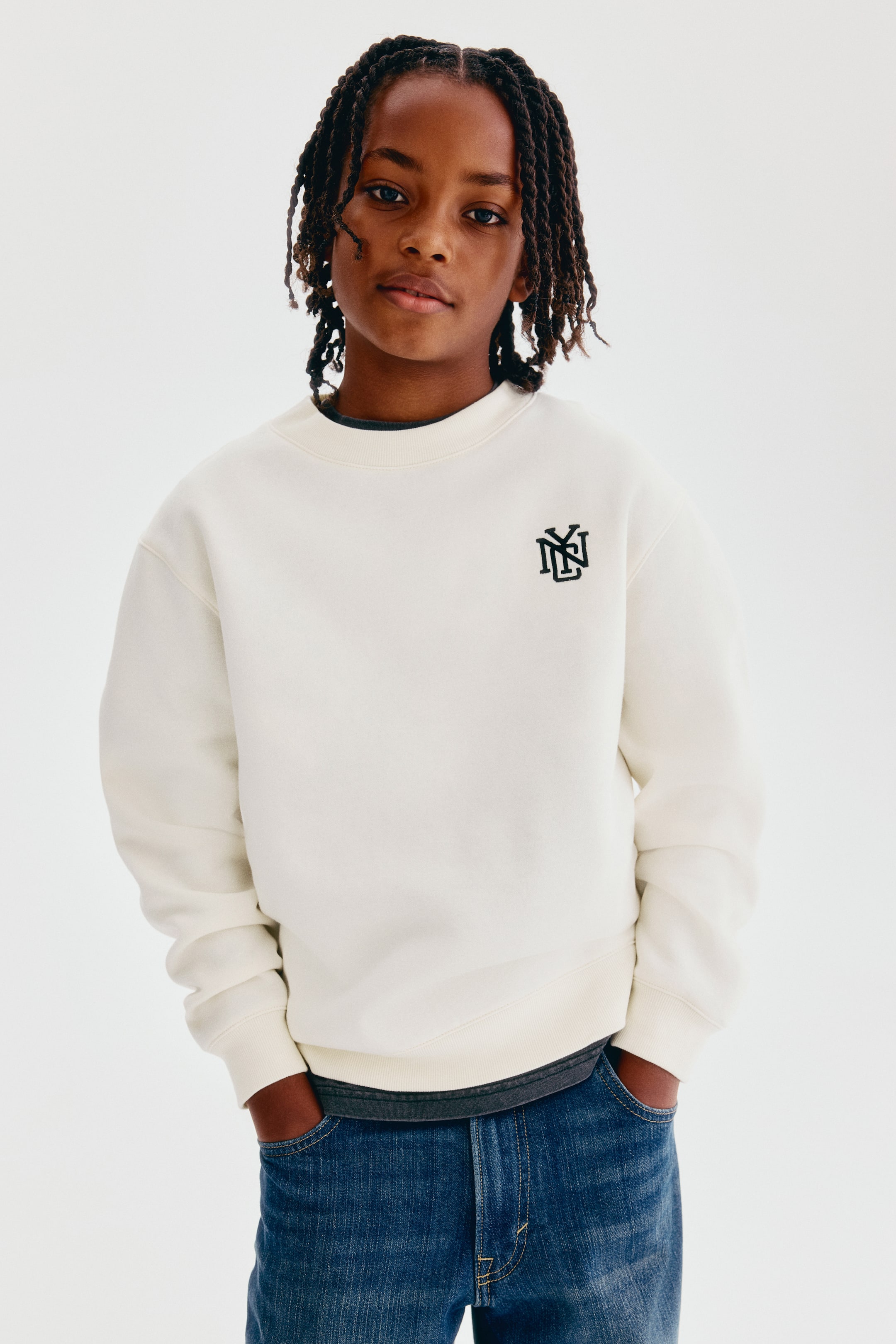 View larger image: A cream-coloured sweatshirt is shown from the front, featuring a round, rib-trimmed neckline, dropped shoulders, and a small black embroidered monogram on the left chest. The sweatshirt has ribbed cuffs and a ribbed hem, and is styled layered over a dark grey T-shirt with blue jeans.