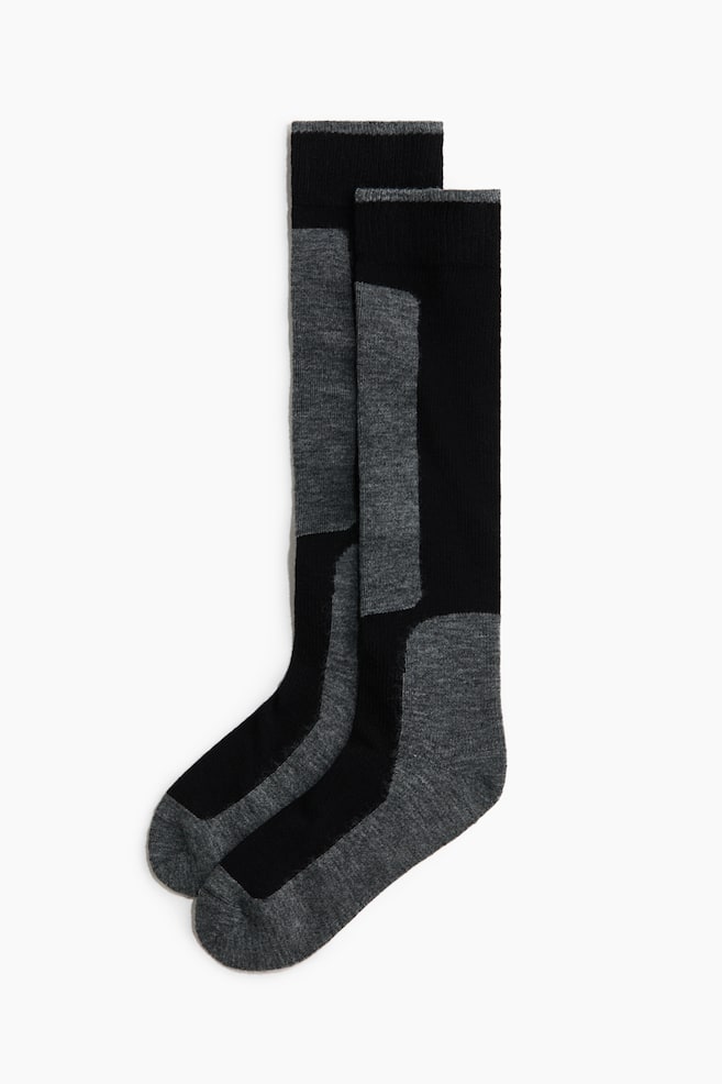 Des chaussettes montantes sont posées à plat et se chevauchent en vue de profil, mettant en valeur un design bicolore en noir et gris chiné. Les chaussettes présentent des bords côtelés avec un fin liseré gris, un empiècement gris le long du tibia, ainsi que des talons et des pointes contrastés gris. - 1