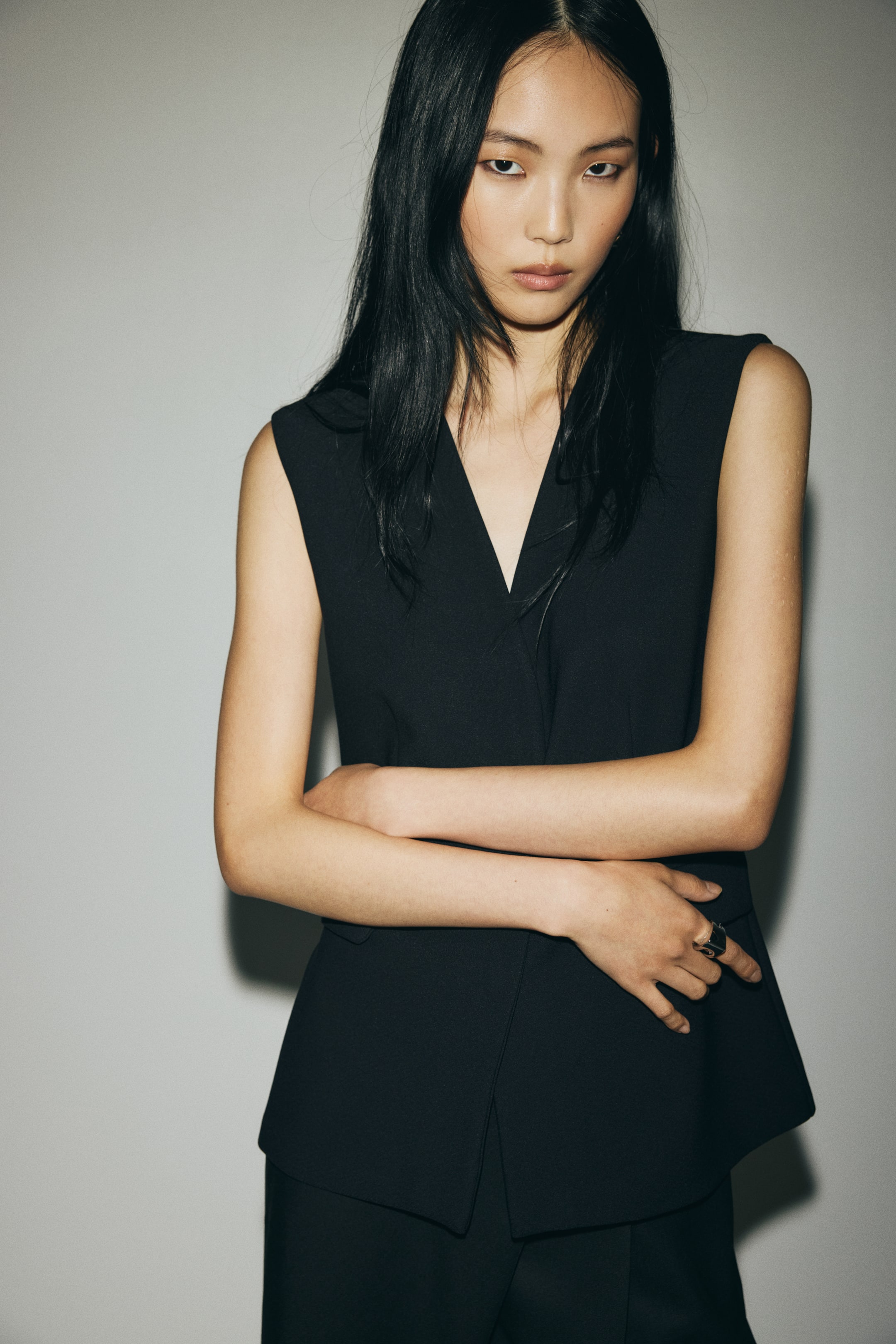 Ver imagen más grande: Un chaleco negro sin mangas presenta un escote en V profundo y un frente estilo envolvente, con un panel superpuesto al otro. El chaleco estructurado presenta un dobladillo ligeramente asimétrico y una sutil abertura lateral en la cadera.