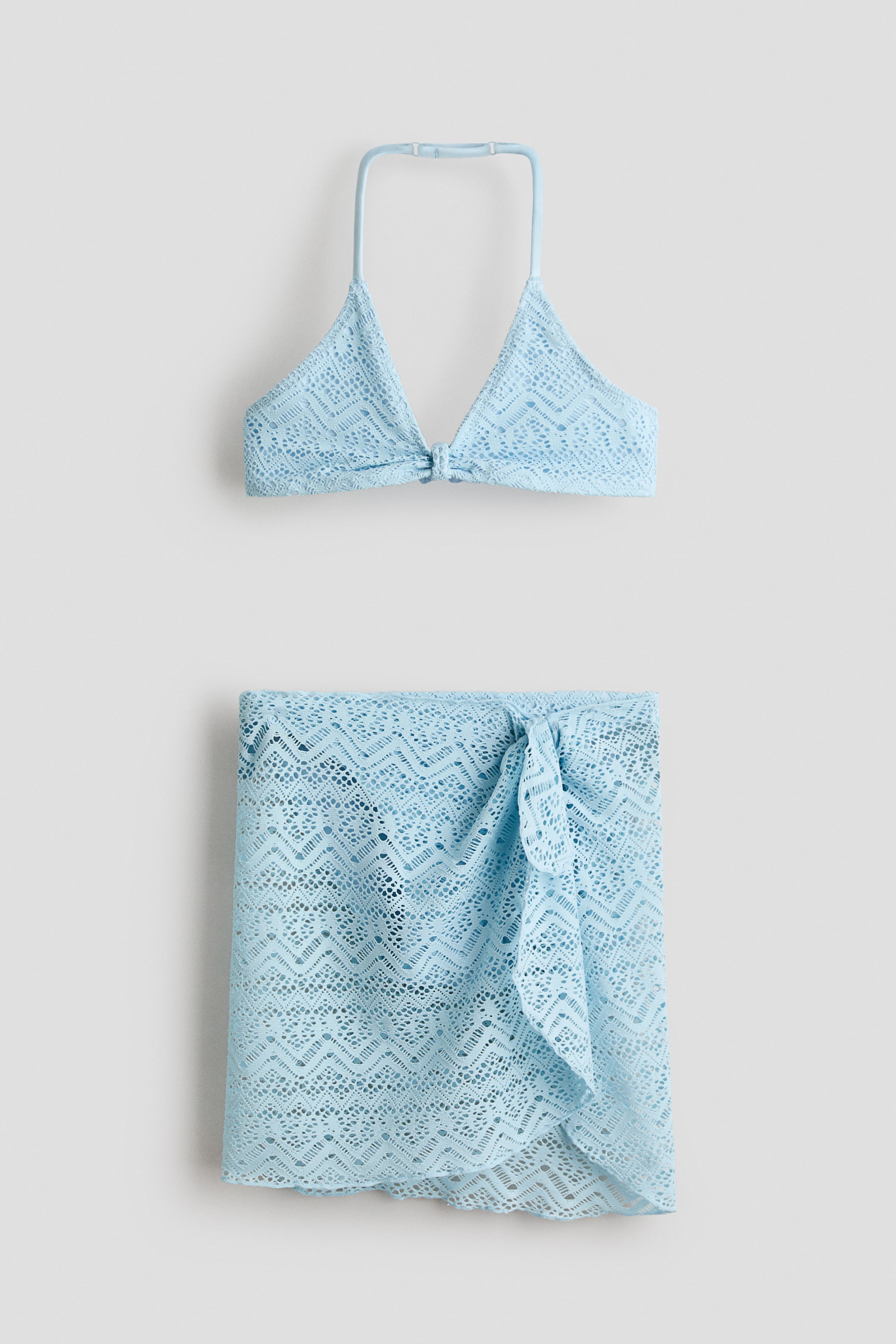 View larger image: A light blue crochet-look bikini set is showcased from the front, featuring a halterneck top and a matching wrap skirt. The fully lined bikini top includes triangle cups with a central knot and a narrow neck strap that has a plastic adjuster, while the unlined skirt displays wide ties fastened in a knot at the front.