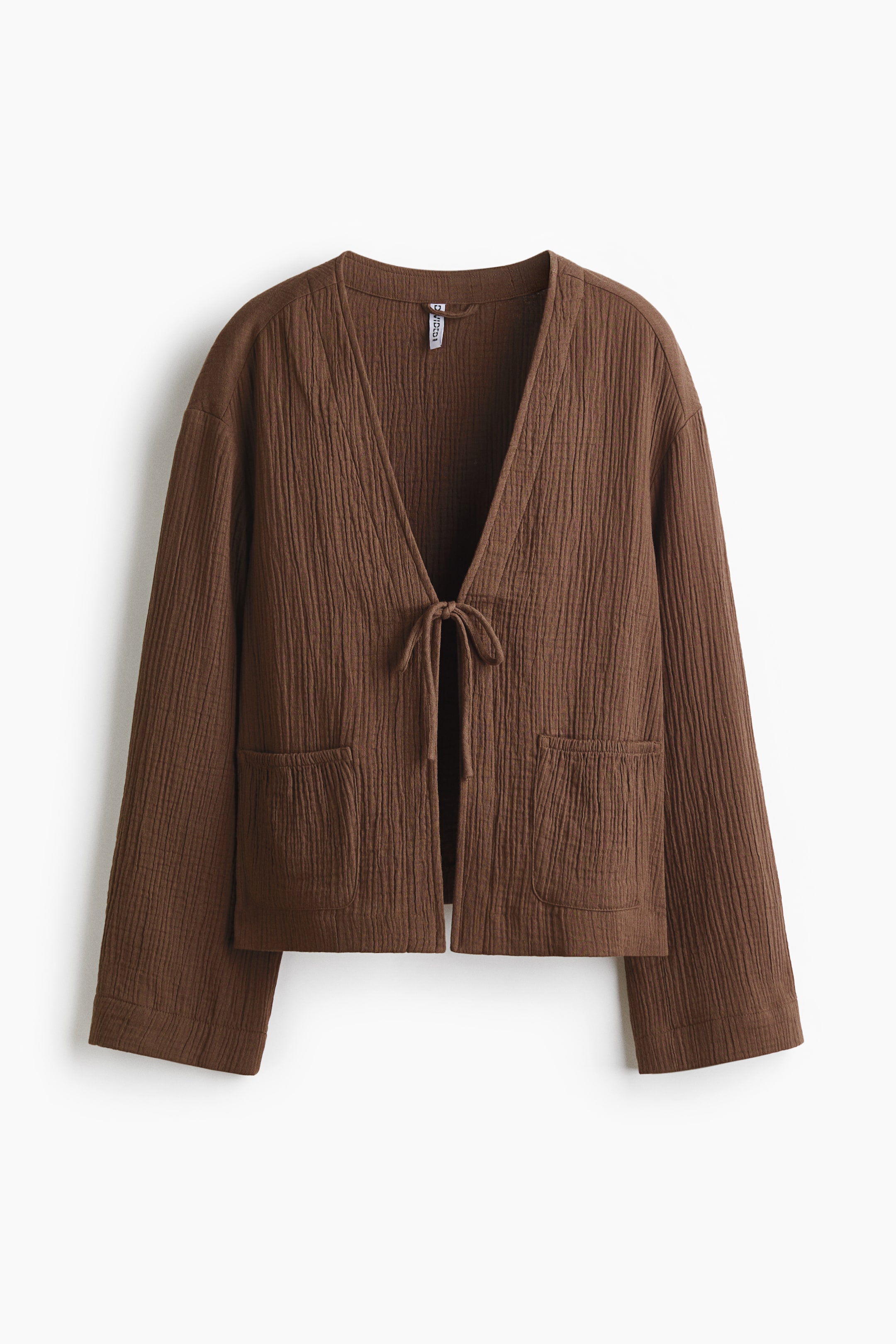 View larger image: A brown cotton muslin jacket is on display from the front, highlighting a deep V-neckline and an open front with slim ties neatly knotted together. It features dropped shoulders and two patch front pockets with narrow elasticated openings.