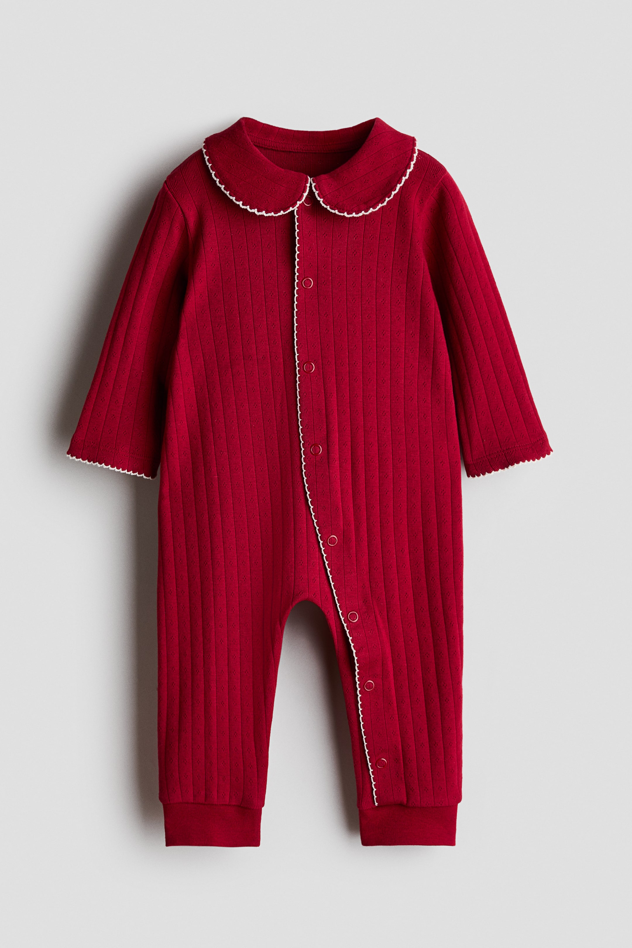View larger image: A deep red pointelle jersey sleepsuit is displayed from the front, featuring a rounded, white lace-trimmed collar, white press studs running down the front and continuing along one leg, long sleeves with picot-trimmed cuffs, and cuffed leg hems.