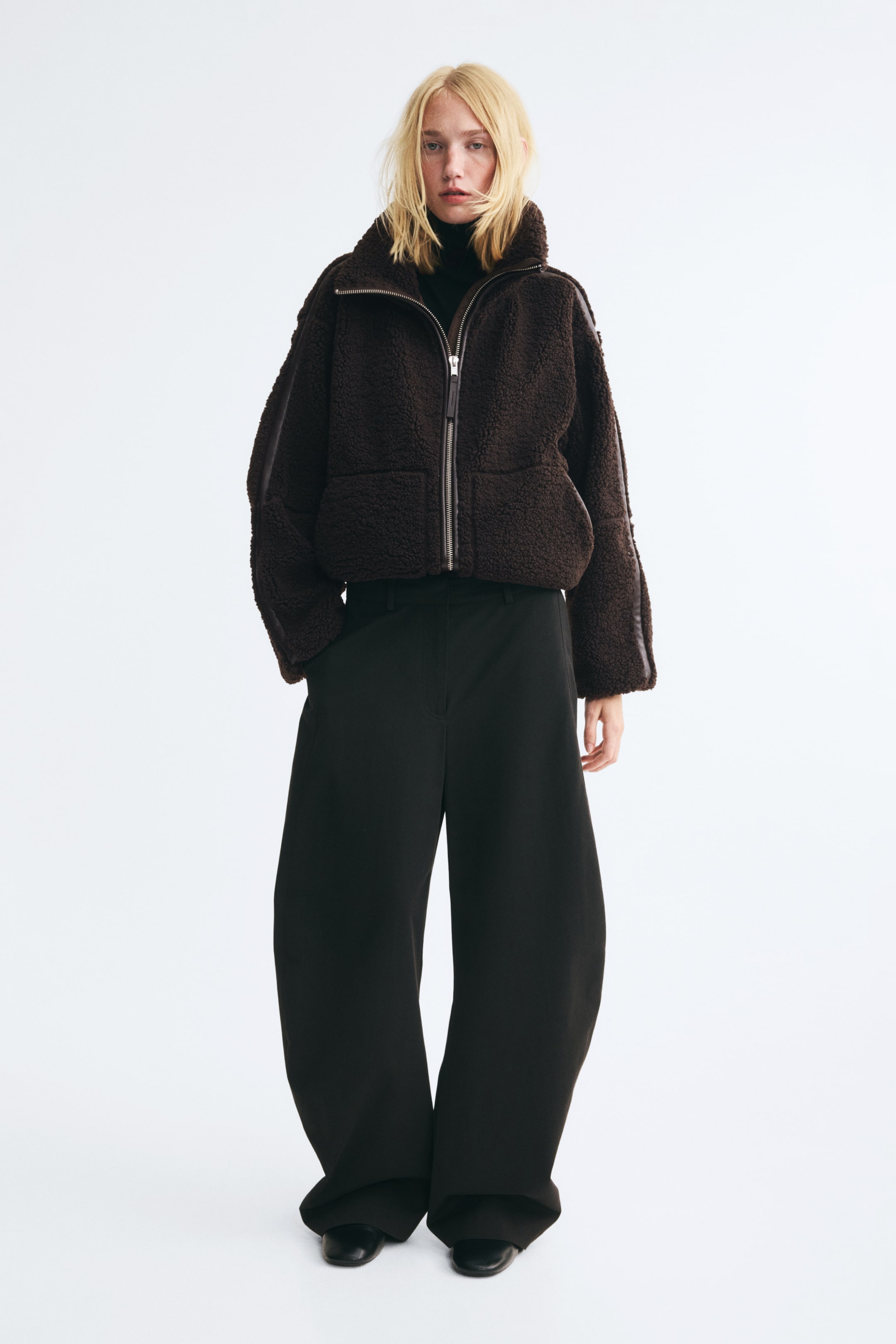 Vis større bilde: En oversized, mørkebrun teddyjakke med funnelkrage, en toveis glidelås i sølv med belagt stoffkant og store sidelommer bæres av modellen. Jakken er stylet over en svart pologenser og kombinert med svarte bukser med vide ben.