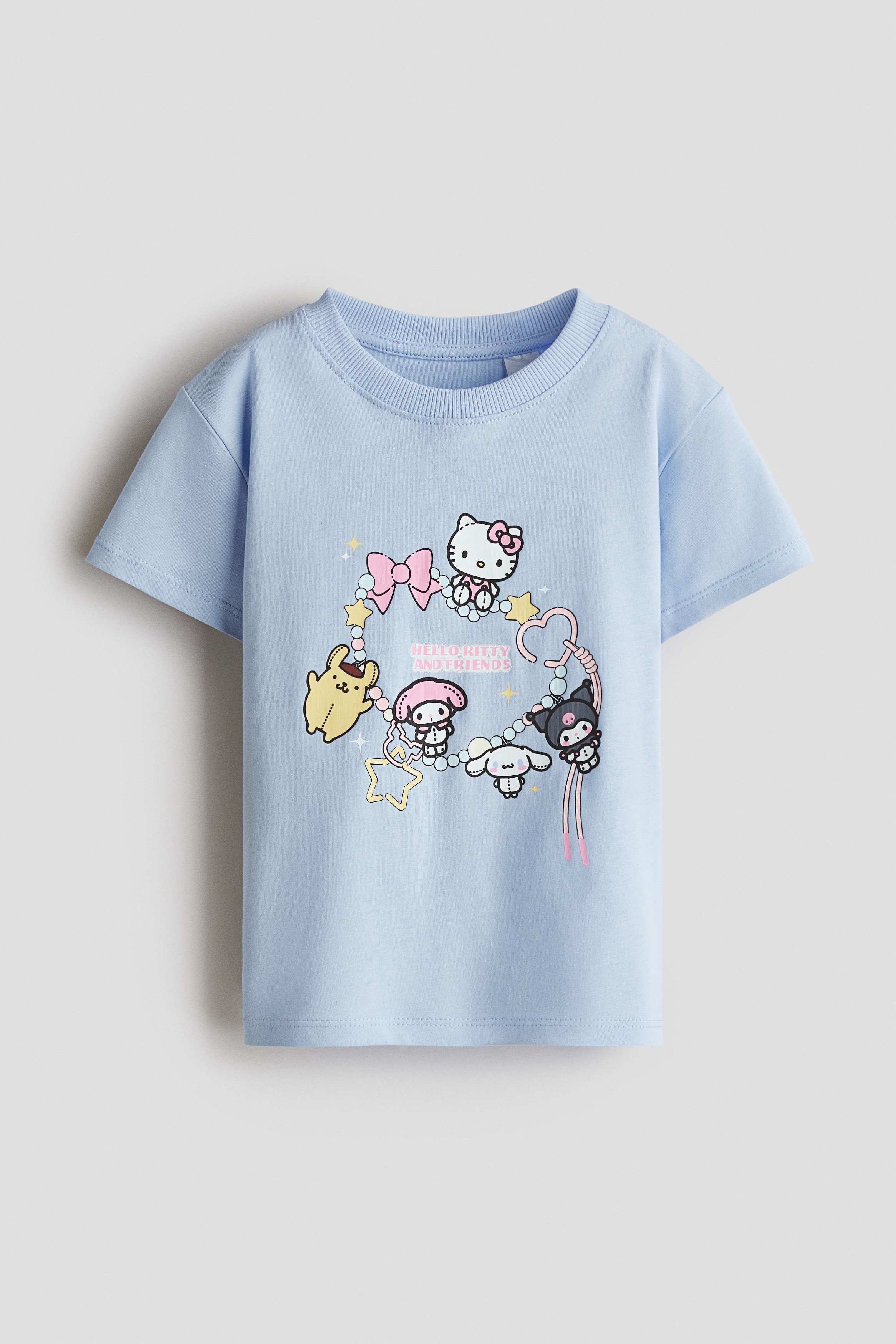 A light blue cotton jersey T-shirt featuring a colourful cartoon print on the front, a round rib-trimmed neckline, and dropped shoulders is worn by the model. The model styles the T-shirt with blue jeans while holding a small, transparent handbag with a pink strap.