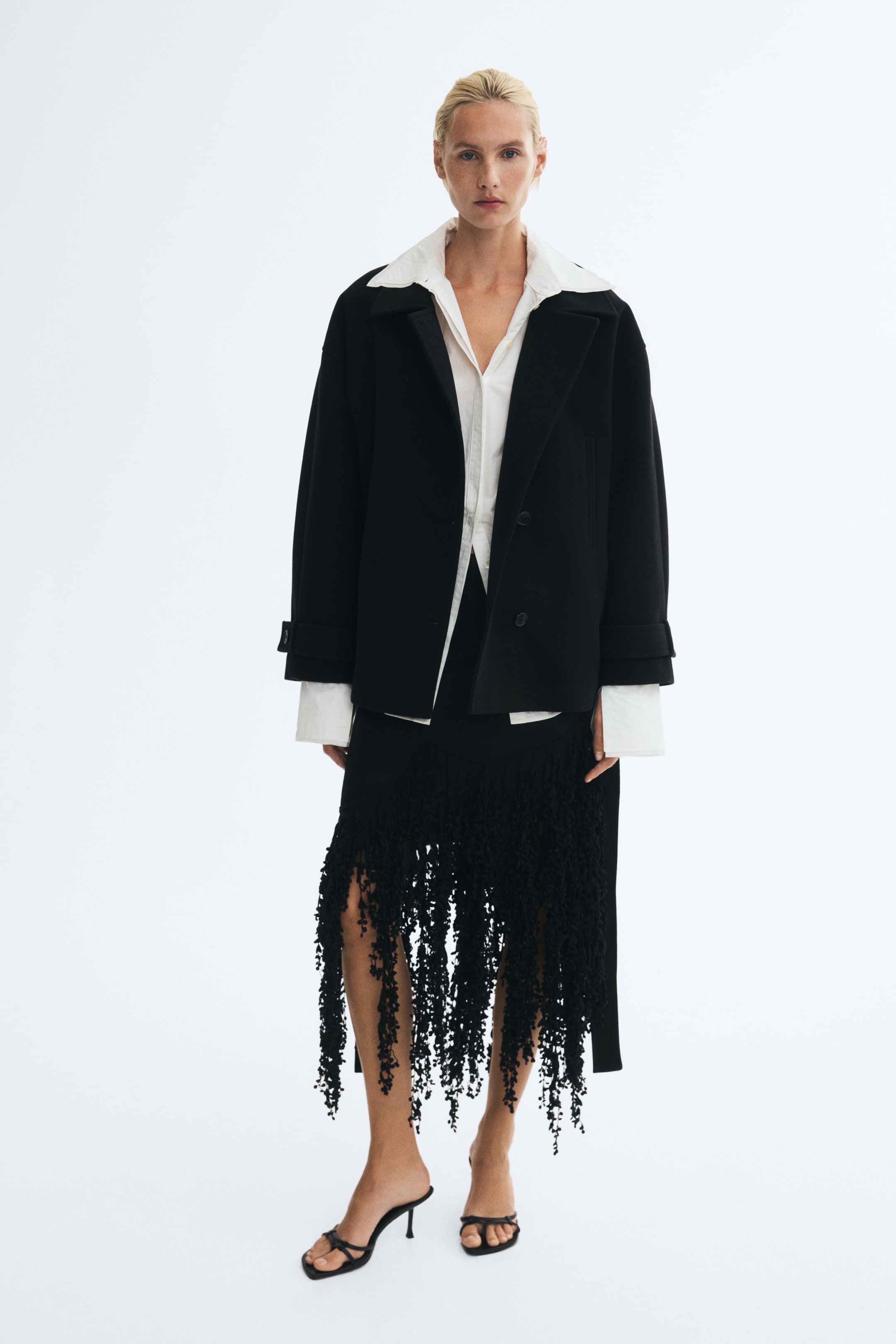 Visualizza immagine più grande: Una giacca in feltro nera con ampio colletto, spalle scese e polsini con alamari è indossata aperta dalla modella. La giacca è abbinata a una camicia bianca con bottoni e a una gonna nera caratterizzata da lunghe frange materiche.
