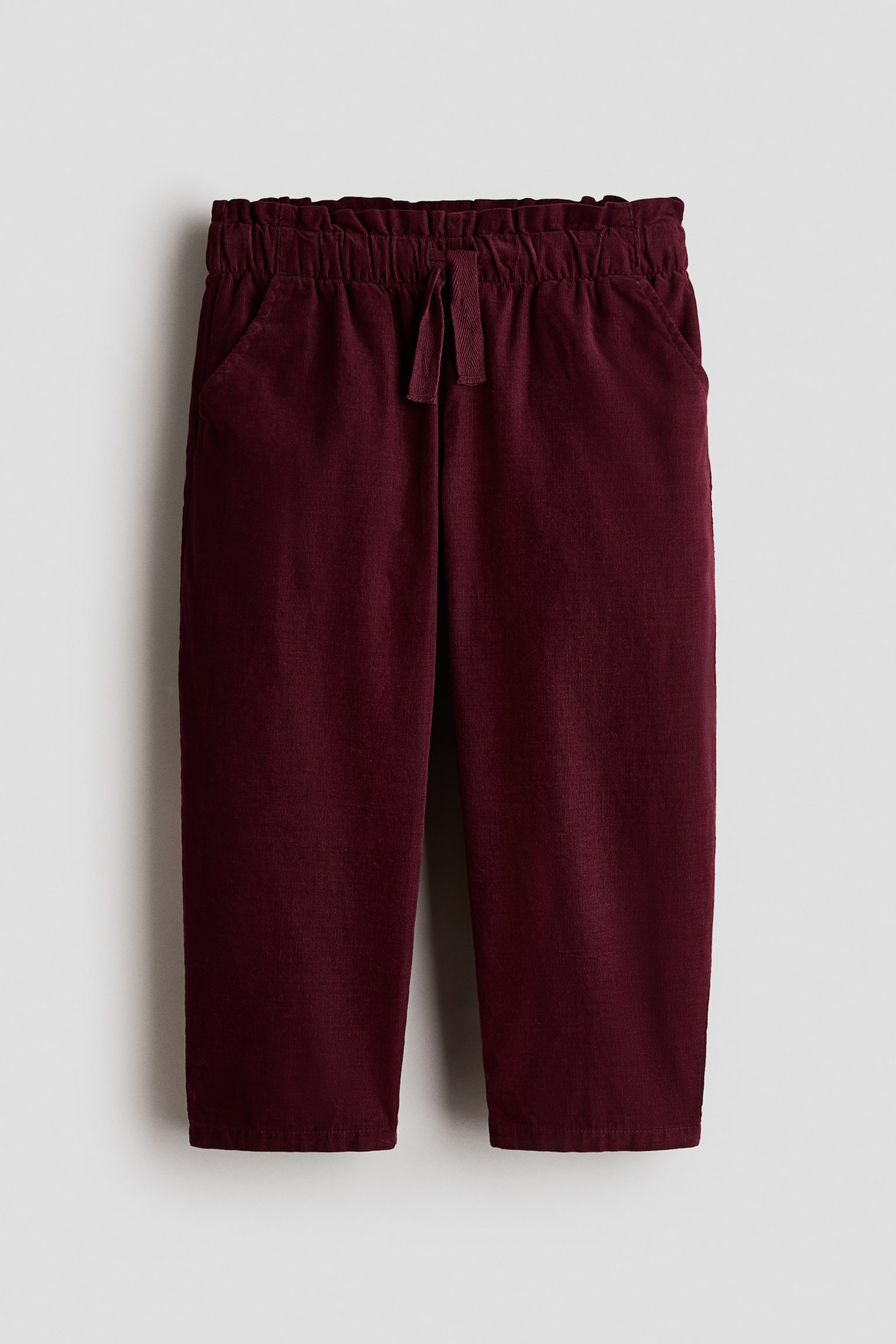 Burgundy corduroy trousers are shown from the front, featuring a ruffled paperbag waistband with a matching fabric drawstring. The trousers showcase side-entry pockets and straight legs.