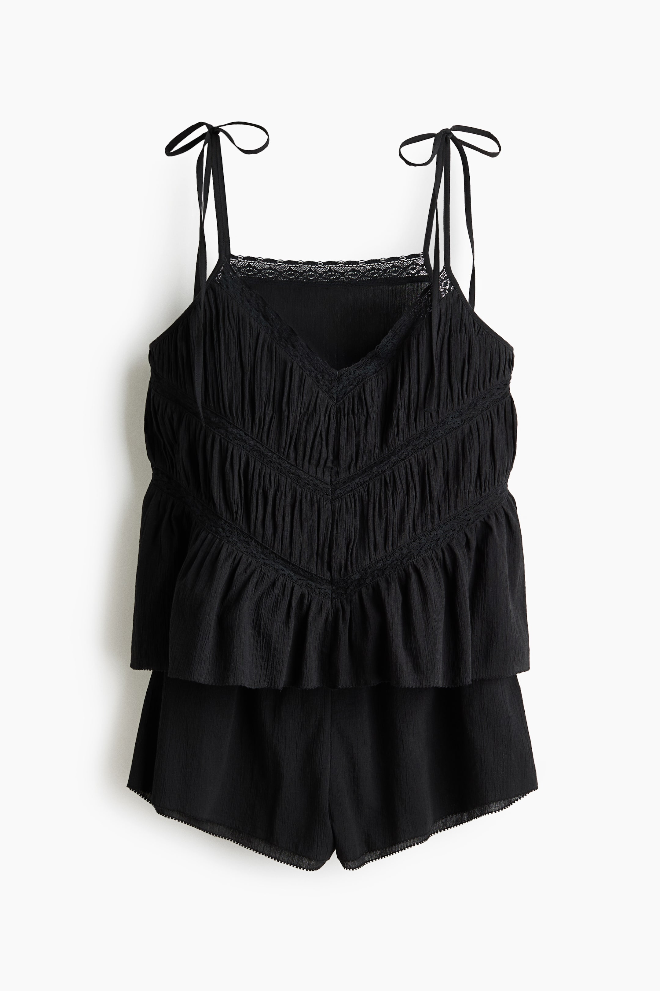 Visa större bild: Ett svart tvådelat set, bestående av ett linne och shorts, visas framifrån. Linnet har en V-ringning med spetskant, knytbara spaghettiband och rynkade paneler som bildar ett V-mönster på framsidan, avslutat med en volangkant, medan de matchande shortsen har en vågig kant.