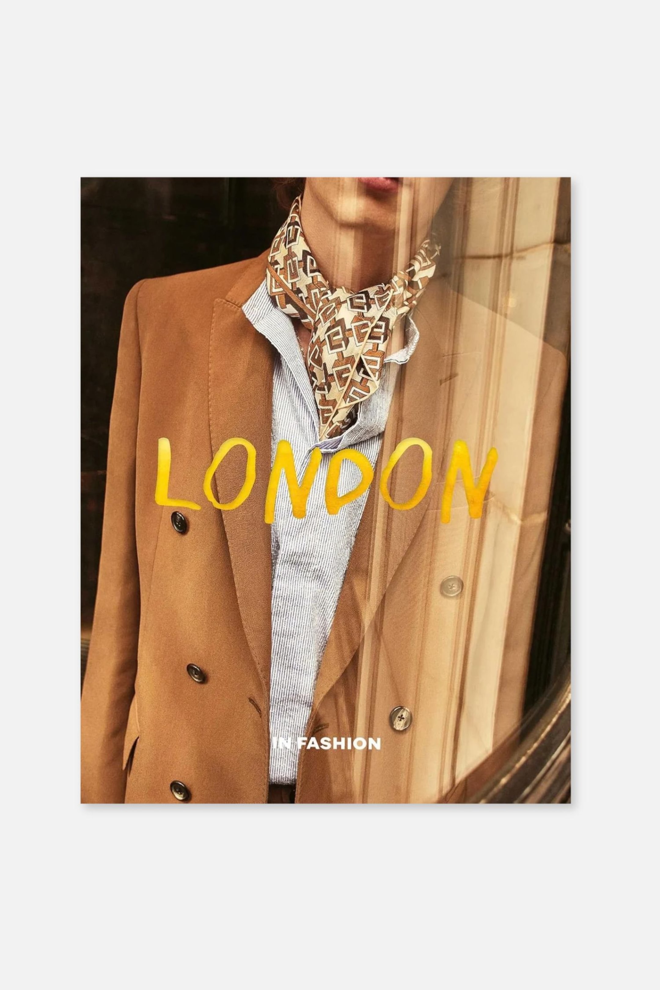 Ver imagen más grande: Un libro titulado «LONDON IN FASHION» presenta en la portada la foto de una persona que lleva un blazer cruzado de color camel, una camisa de rayas azul claro y un pañuelo de seda estampado al cuello. La palabra «LONDON» se extiende por el centro en tipografía de pincelada de color amarillo brillante, mientras que «IN FASHION» aparece debajo en texto blanco en mayúsculas.