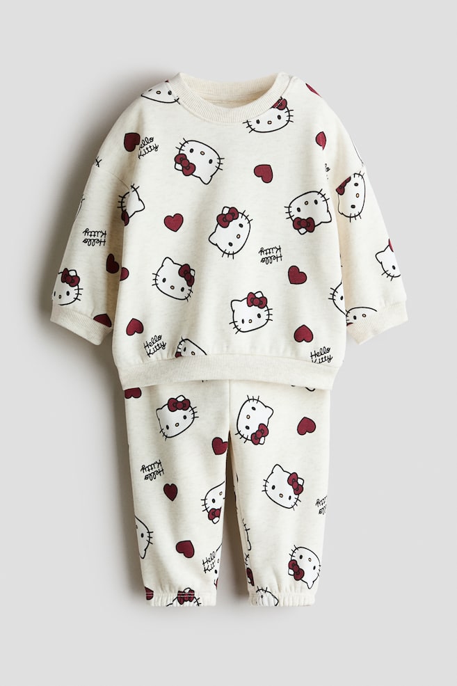A two-piece printed sweat fabric set in cream is shown from the front, featuring an all-over print of Hello Kitty faces, red bows, red hearts, and text. The set consists of a long-sleeved crewneck sweatshirt with ribbed trim and matching sweatpants with elasticised ankle cuffs. - 1